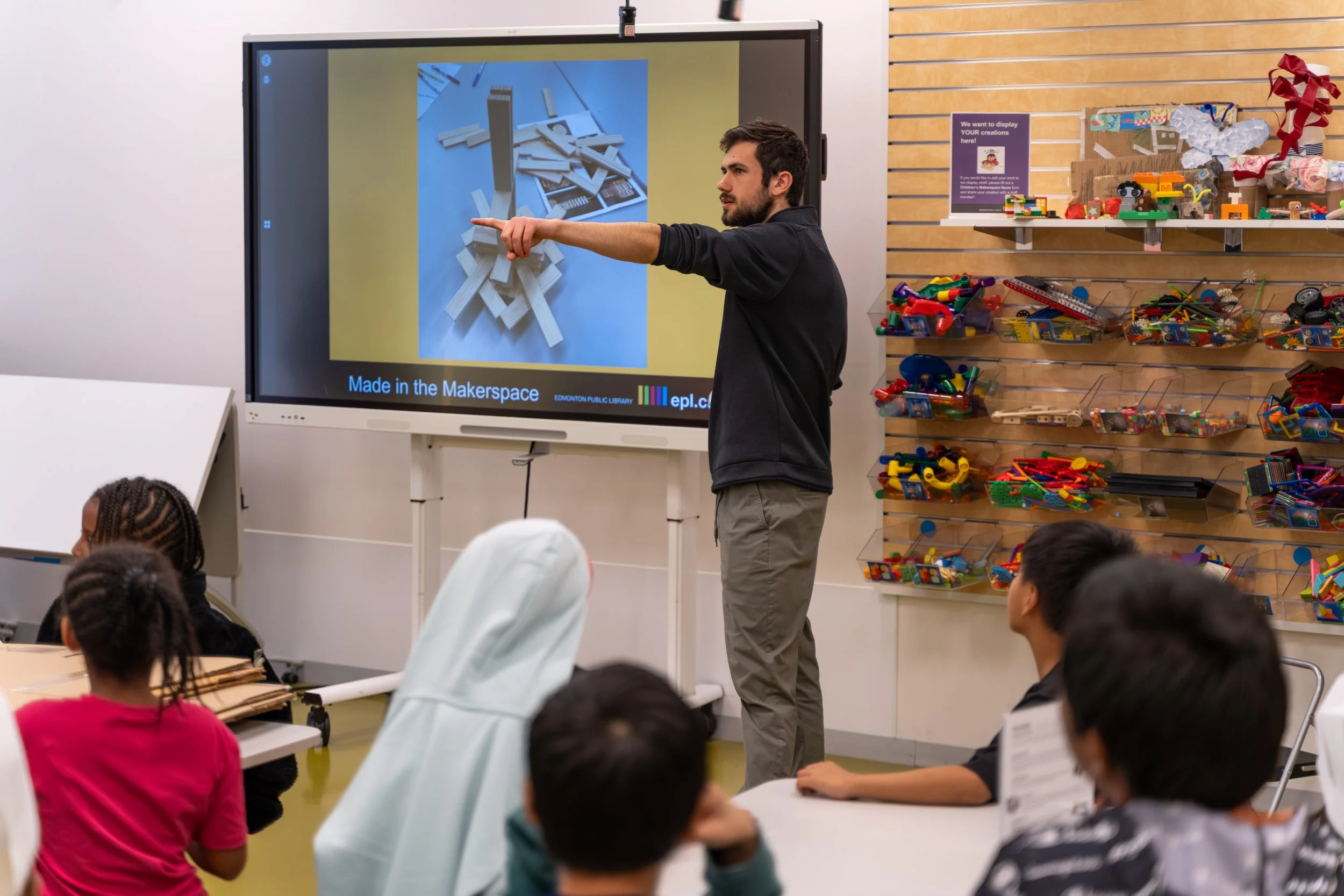 A man is giving a presentation to children in a classroom, pointing at a digital screen displaying a photo of wooden building blocks. The classroom has shelves with various toys and craft supplies.