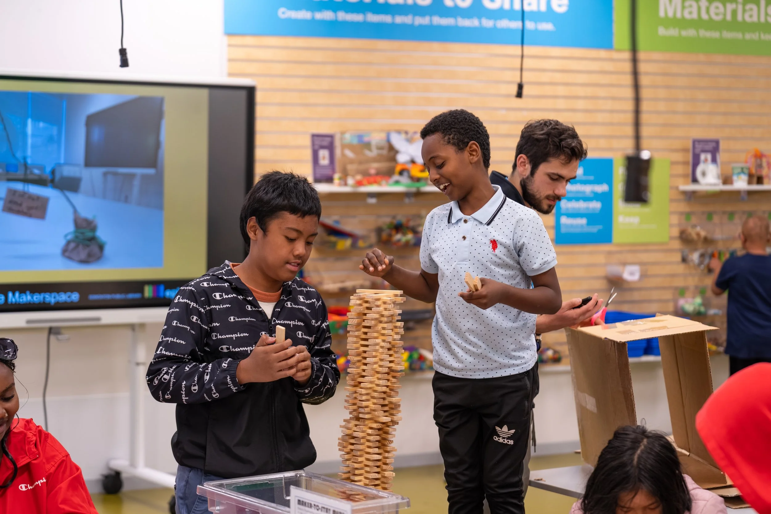 Three children play a giant Jenga game at a community event. They are smiling and appear engaged in the game, with the background showing a brick wall and colorful signs.