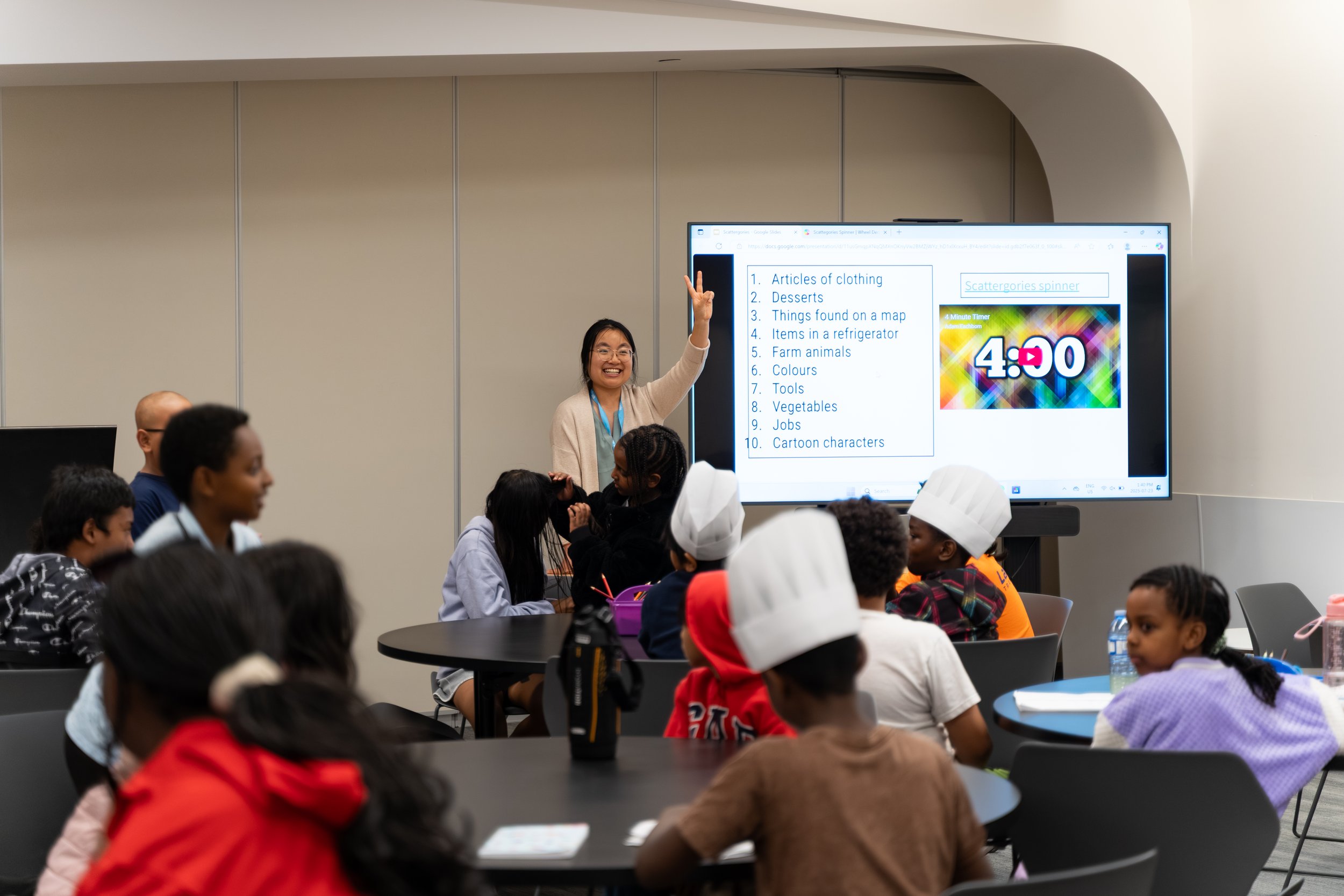 A diverse group of children and a teacher in a classroom, with the teacher standing next to a large screen displaying a list of categories and a timer. The children are sitting at tables, some wearing chef hats.