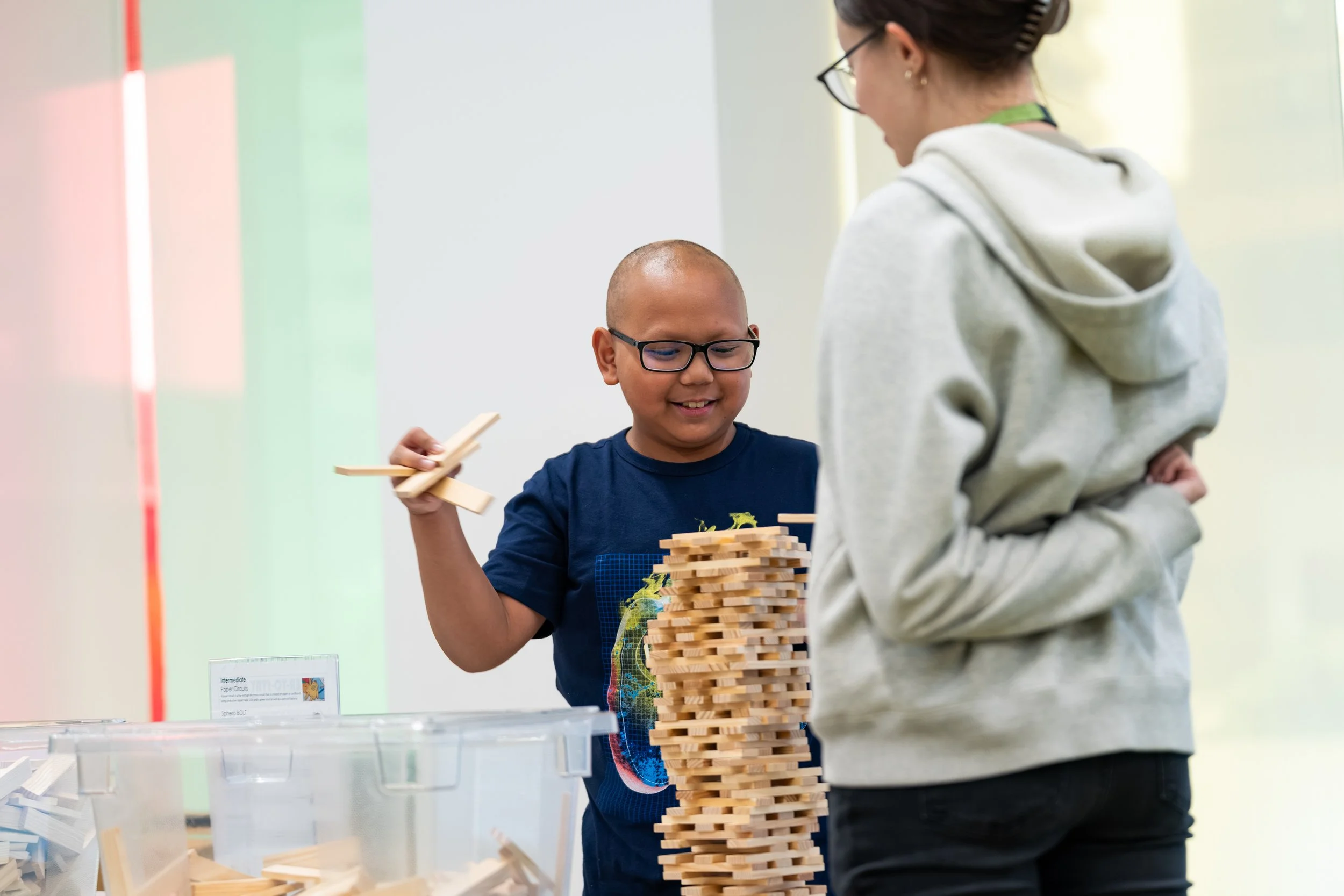 A boy building a tower with wooden blocks at an indoor event while a woman, possibly a caregiver or teacher, watches nearby.