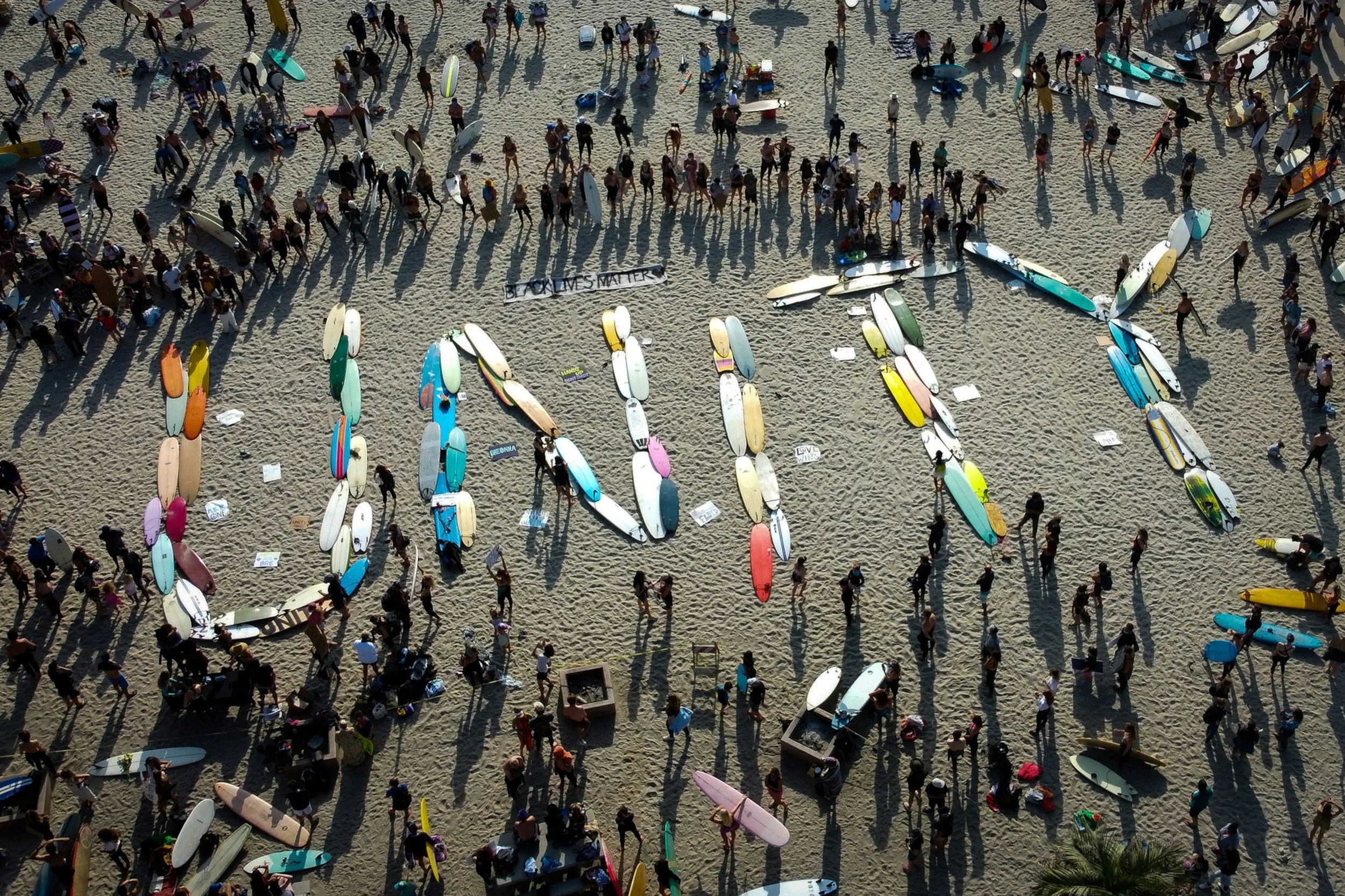Aerial view of a crowded beach with numerous people and colorful surfboards scattered on the sand, some people holding surfboards and others walking or standing, with a message that reads 'Black Lives Matter' on a banner.