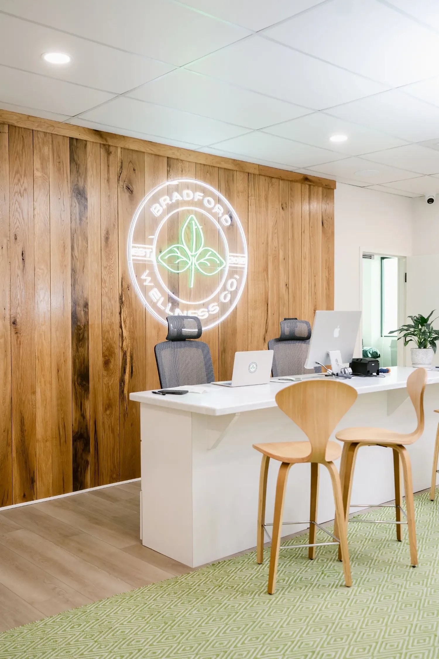Office reception area with a wooden wall, white desk with two laptops, gray chairs, and a neon sign reading 'Bradford Wellness Co.' featuring a green leaf logo. There is a plant near a window and a green patterned rug.
