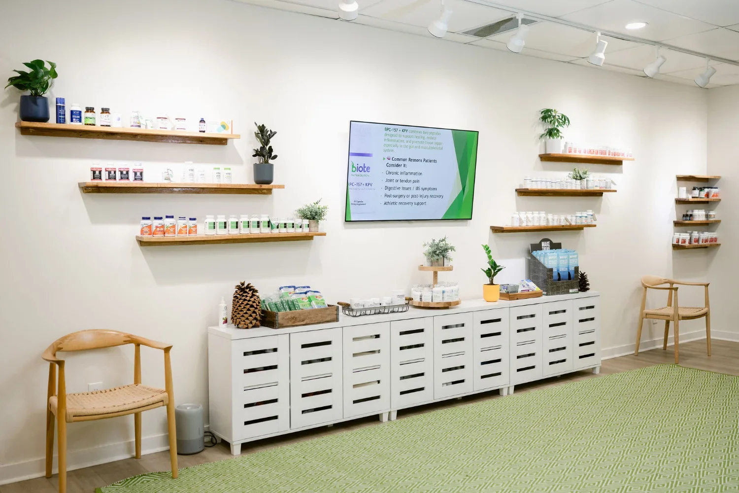 Display of health supplements and natural products in a wellness clinic or pharmacy with shelves on a white wall, a large monitor screen, and wooden chairs.