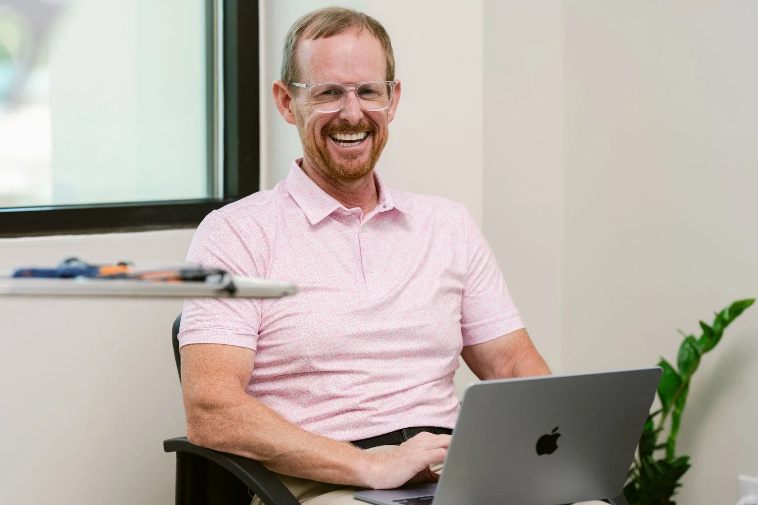 A man with glasses and a beard smiling while sitting at a desk with a silver MacBook in front of him, in a bright office with a window and a plant in the background.