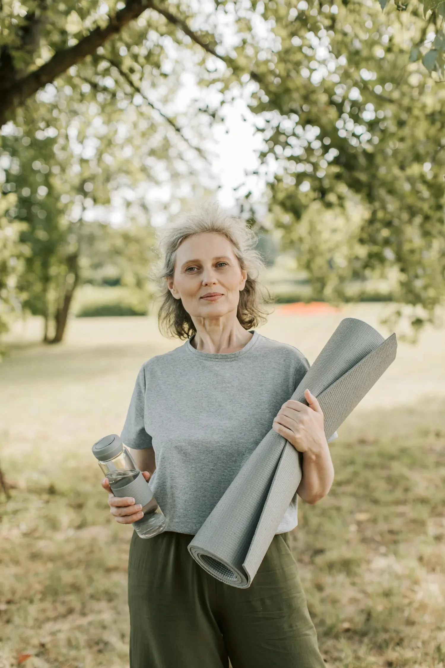 An older woman outdoors holding a water bottle and yoga mat, wearing a gray t-shirt and green pants, with trees and sunlight in the background.
