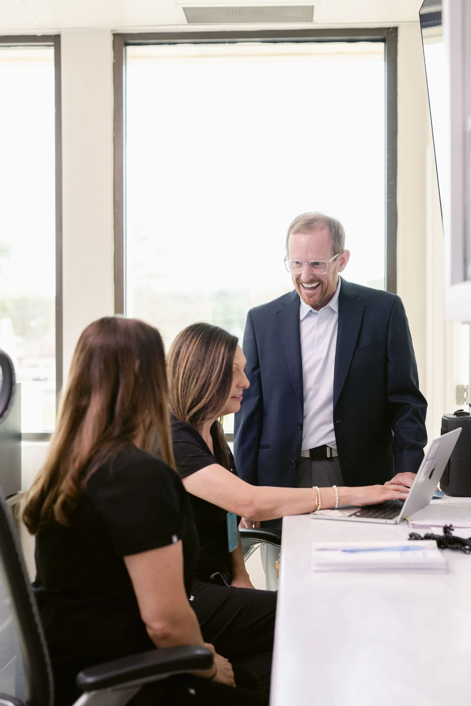 Dr Chad Bradford in a dark suit and glasses is smiling and talking to a group of women seated at a table, working on a laptop, in a bright office with large windows.