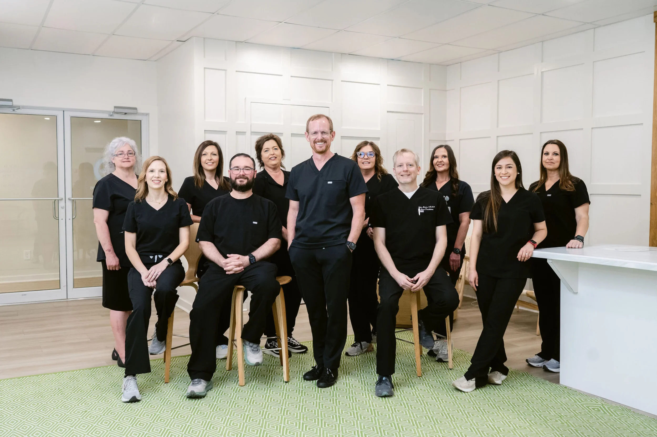 A group of ten healthcare professionals, including men and women of diverse ages and ethnicities, posing together in a medical facility with white walls and wooden flooring.