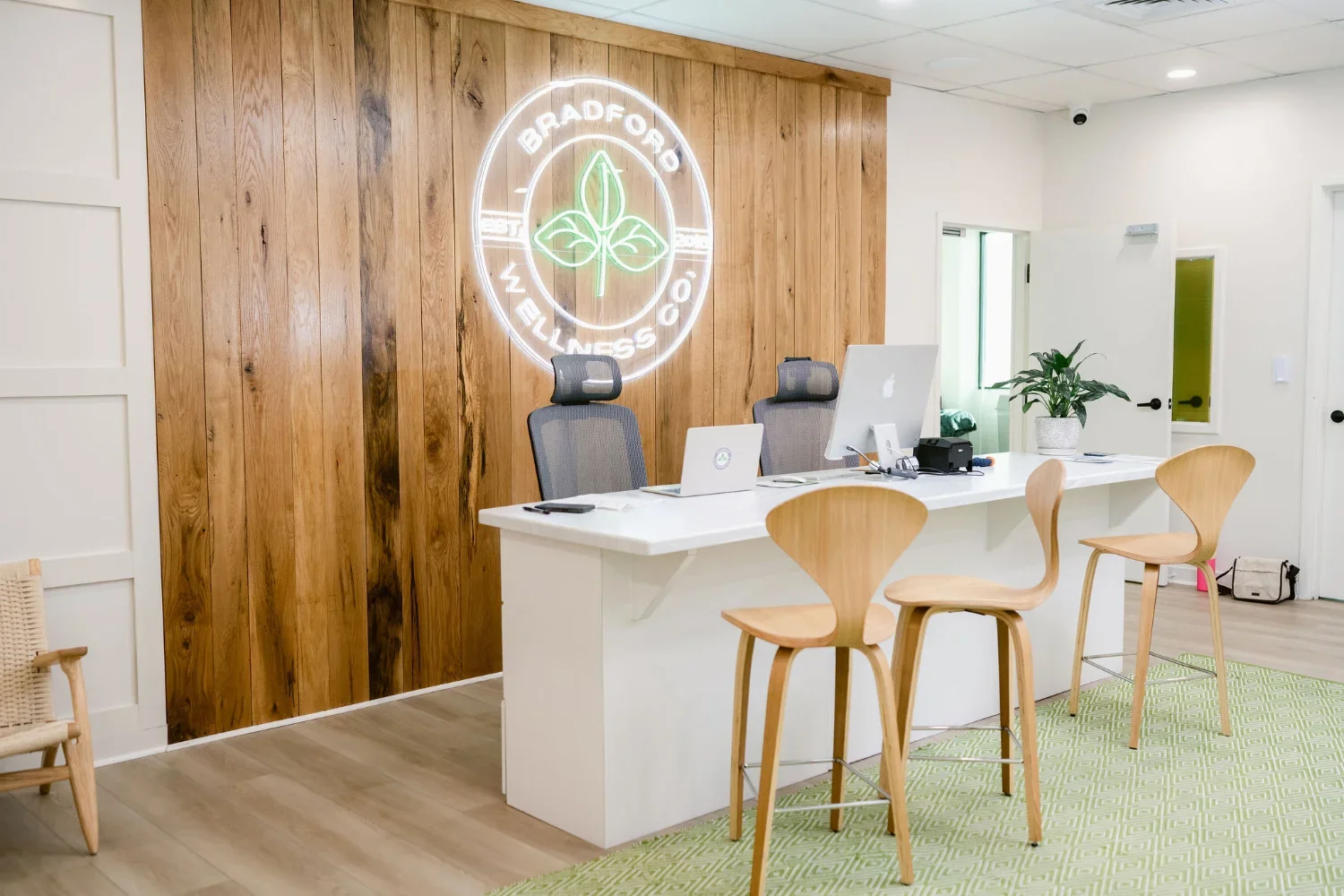 A modern medical office reception area with a white desk, two ergonomic chairs behind it, and two wooden chairs in front.
