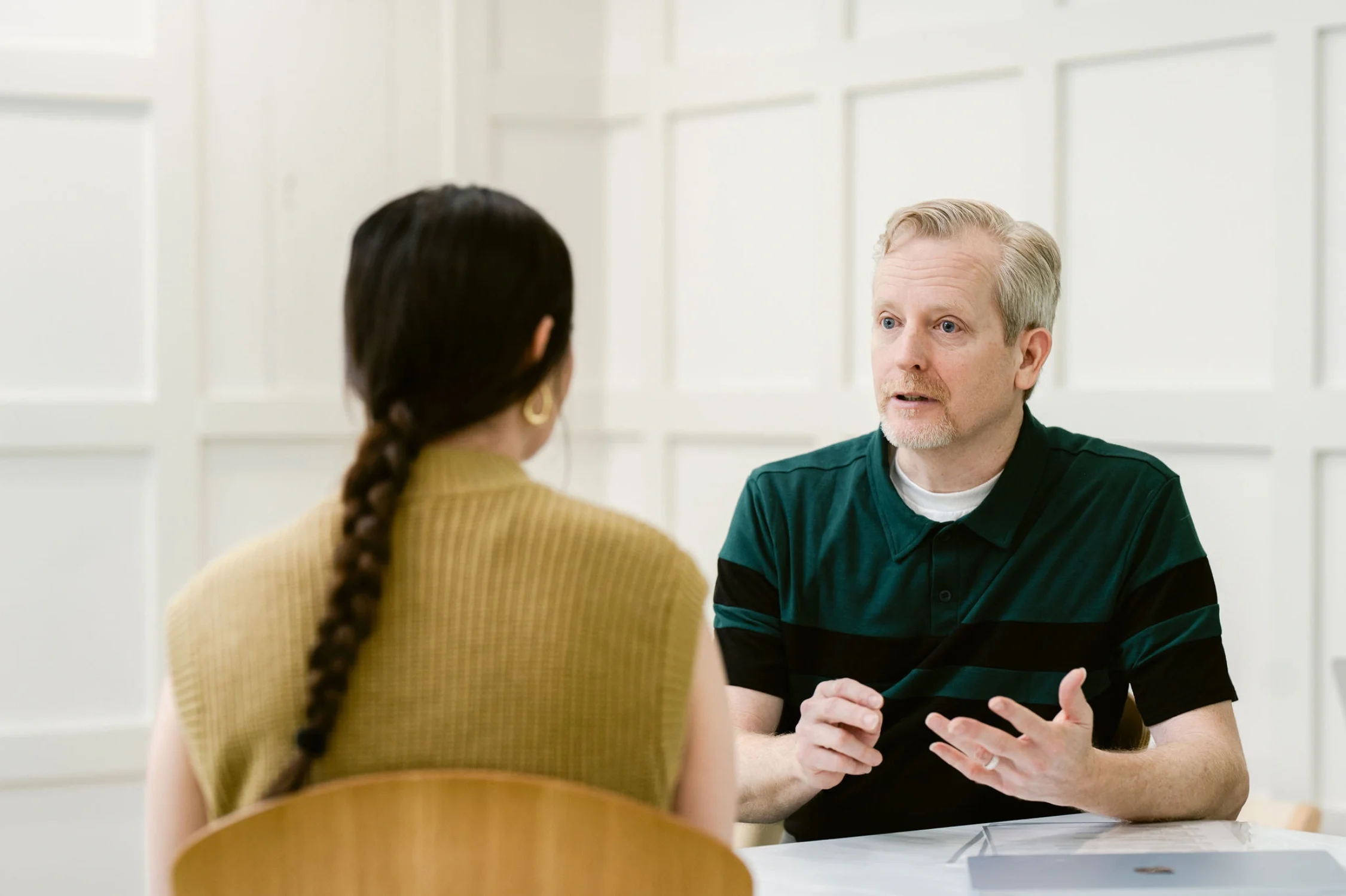A man and woman having a serious conversation in a bright room with white paneled walls. The woman has dark hair in a braid and the man has blond hair, wears a green and black striped shirt, and appears to be speaking