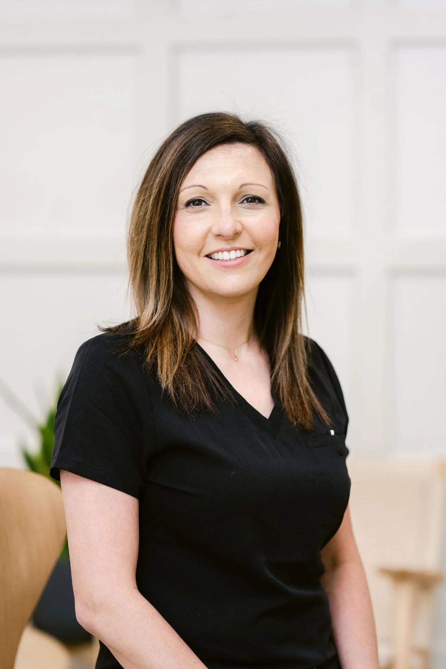 A woman with long brown hair smiling, wearing a black top, sitting in a room with white walls and a plant in the background.