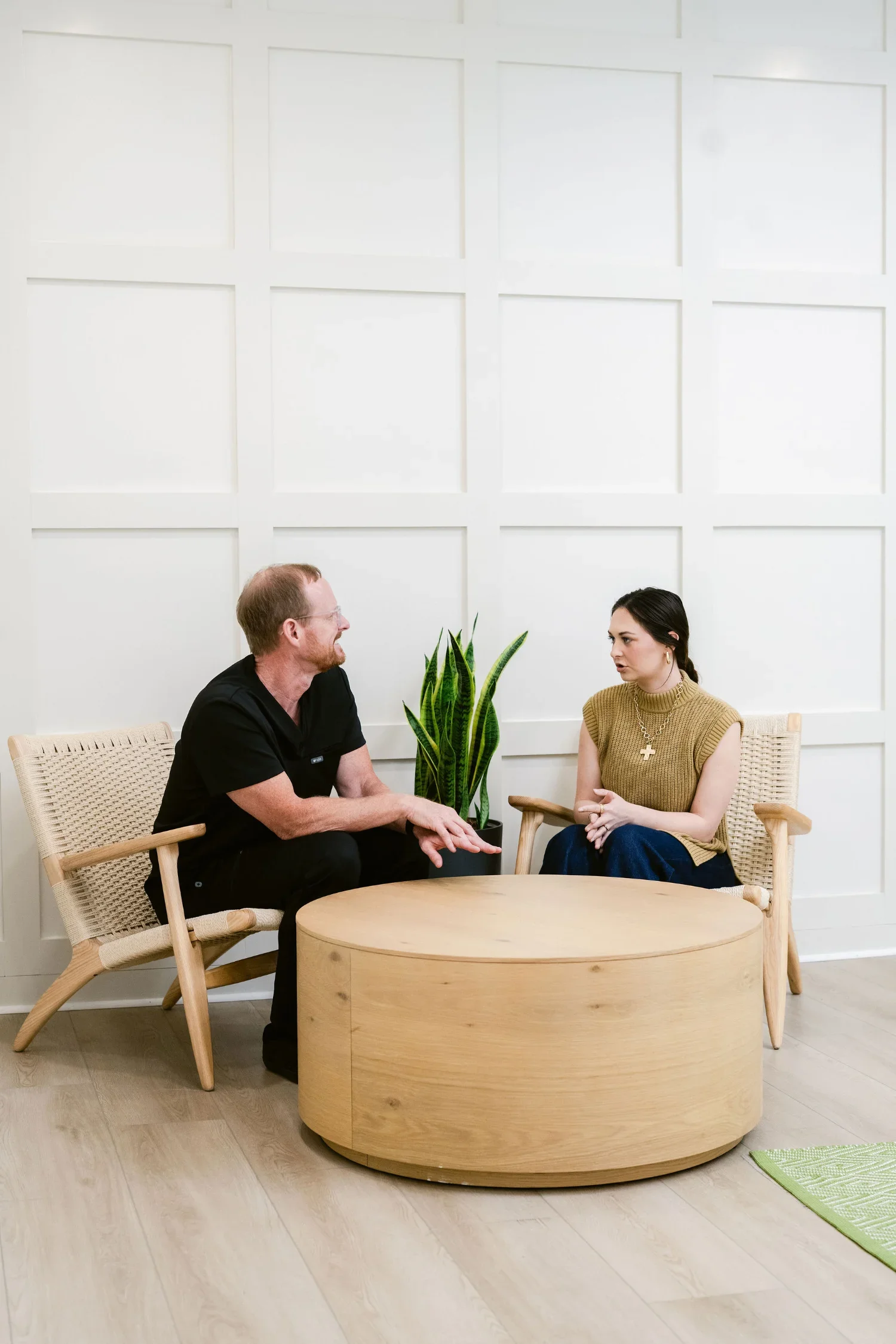 Two people are sitting and talking at a round wooden table in a bright, minimalistic room with a white paneled wall. There is a large green plant between them, and a light-colored woven chair on each side of the table.