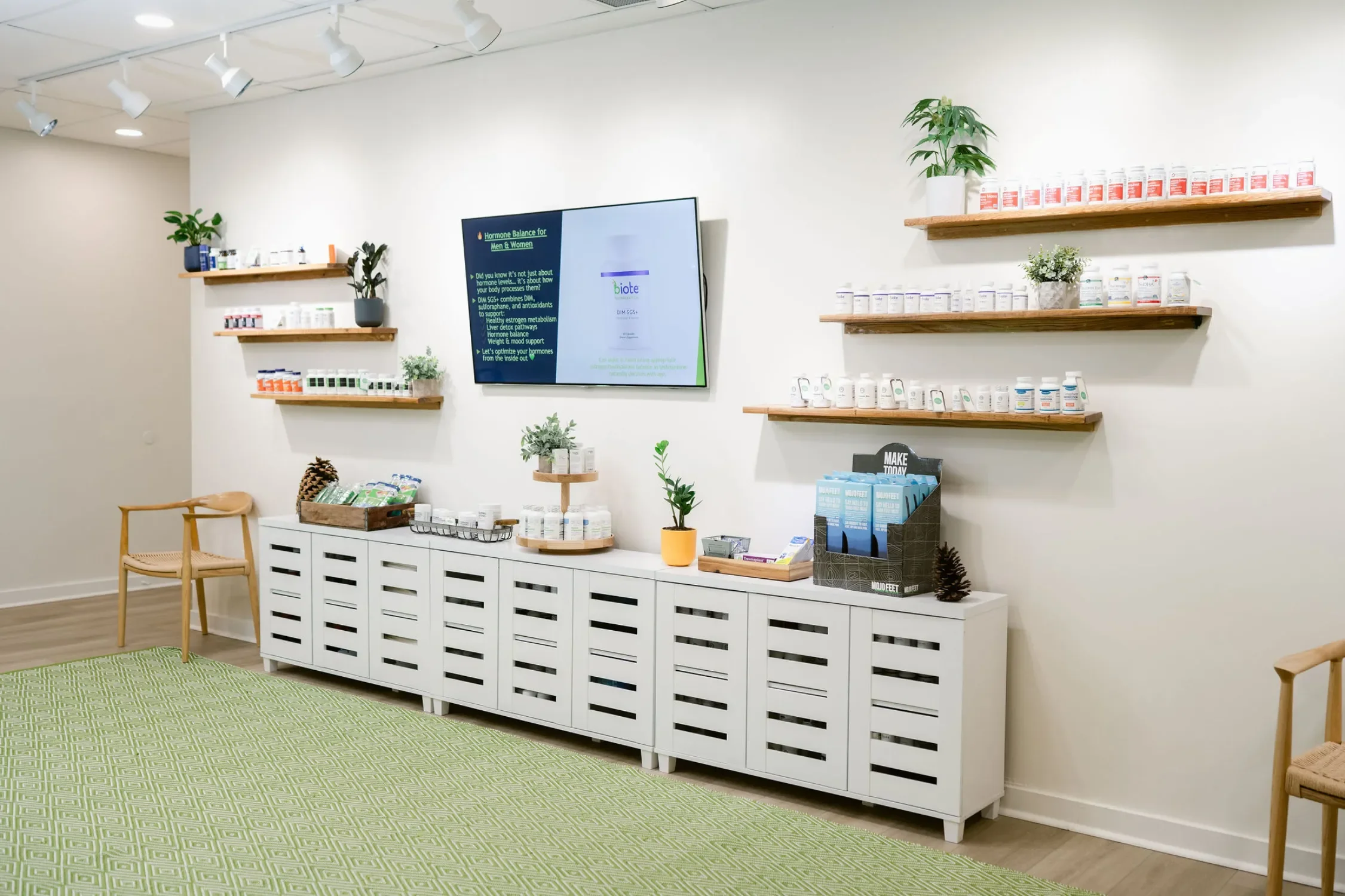 A wellness or supplement store with white walls, wooden shelves displaying bottles and containers, a digital screen on the wall, and green plants.