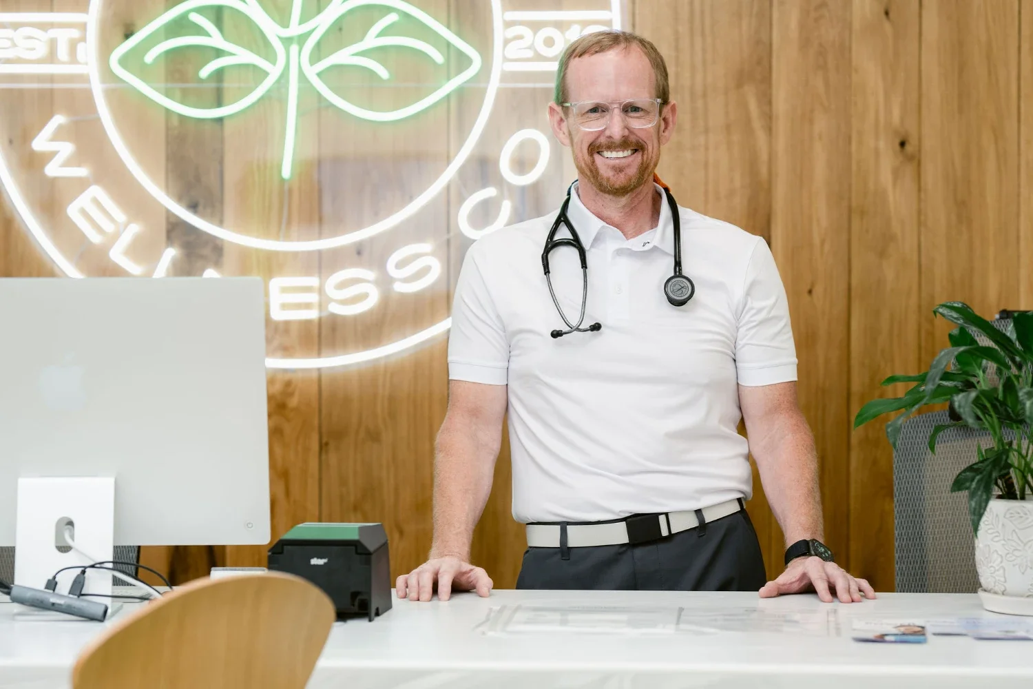 A smiling male doctor with a stethoscope around his neck, standing at a white desk with a computer and office supplies, in a modern office with wooden wall panels and a neon sign in the background.