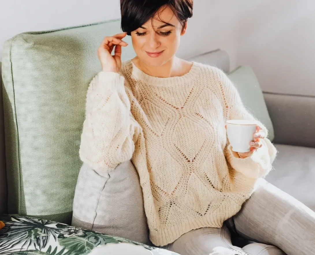 Woman sitting on a light green sofa, holding a white mug, wearing a cream-colored knitted sweater, with short dark hair, smiling gently