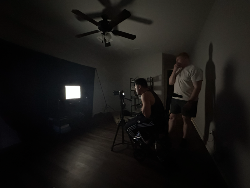 Two men in a dark room with a ceiling fan, one sitting on a chair with a camera, recording a CRT TV screen for "Static", and the other standing. 