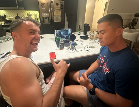 Two men sit at a kitchen counter with voice recording equipment, including microphones, a laptop, and a phone placed on the counter. 