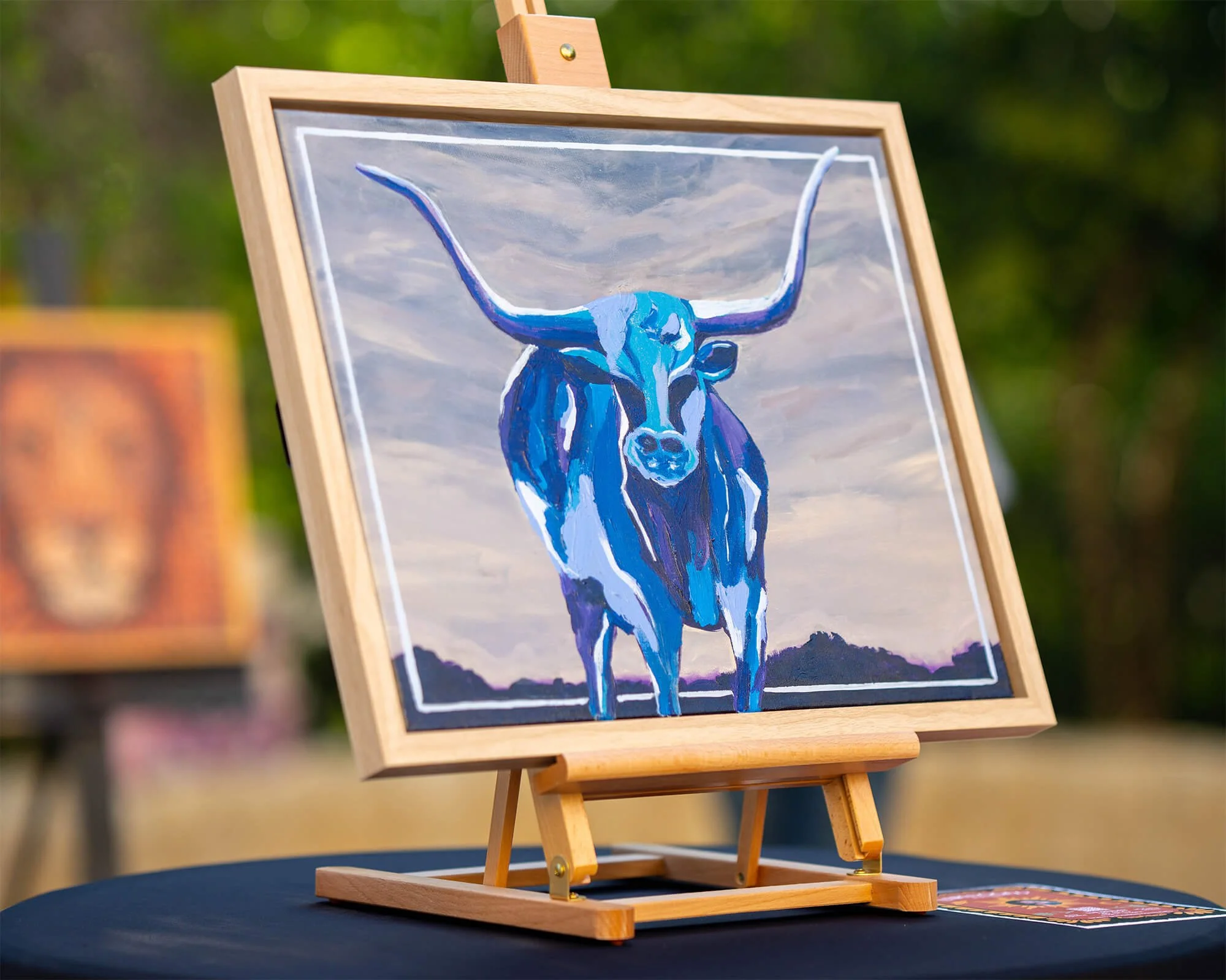 Blue longhorn painting displayed on an easel at an outdoor art exhibition