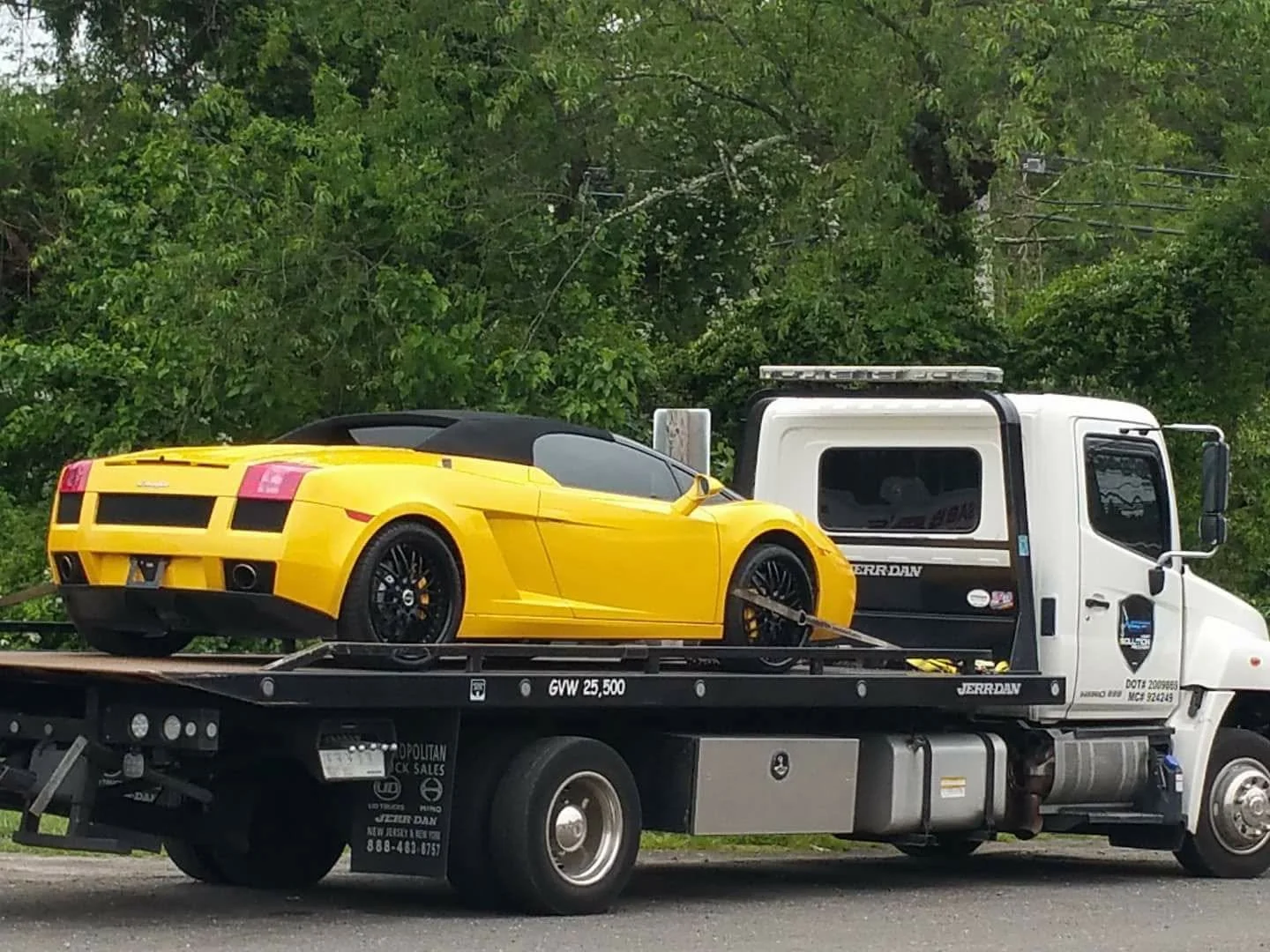 Yellow sports car being transported on a flatbed tow truck with a background of green trees.