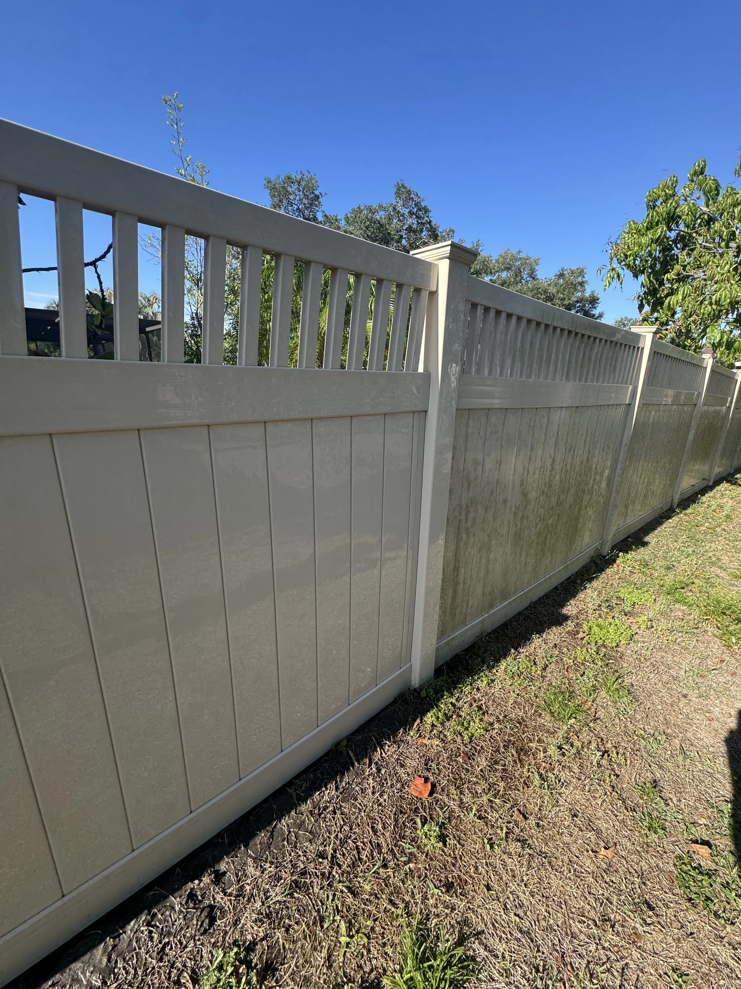 A beige vinyl fence with a lattice top runs along a grassy area with some weeds and small plants, under a clear blue sky. has dirty algae on one side. before and After cleaning of Fence