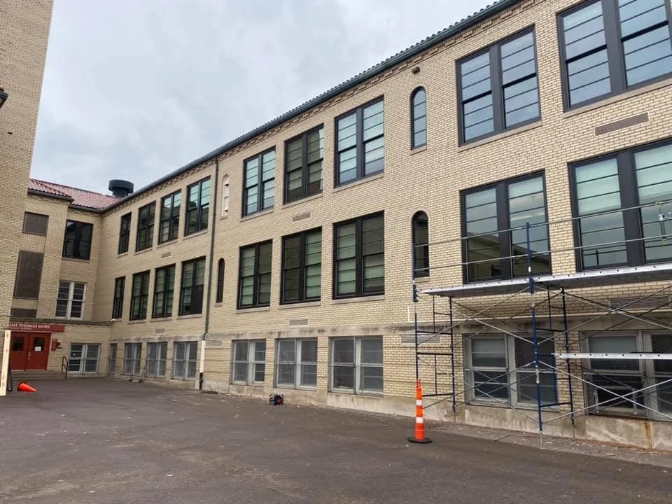 Construction scaffolding outside a multi-story brick building with large windows, on a paved area with traffic cones.