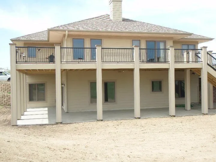 Back view of a two-story house with a porch or balcony on the upper level and a concrete area on the ground level, supported by wooden columns, with stairs on the right side leading up to the balcony.