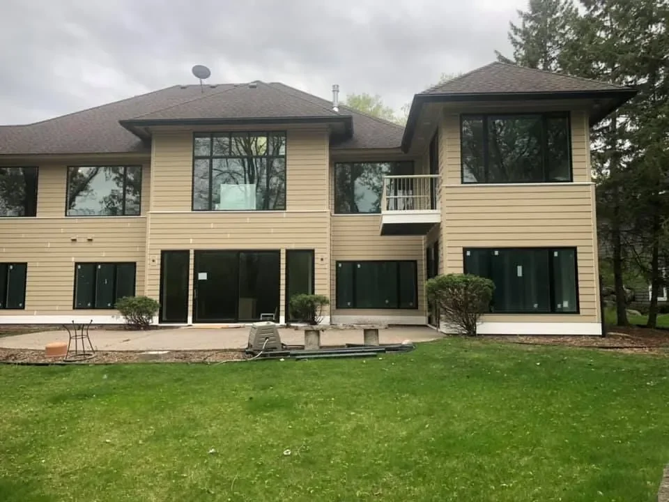 A two-story house with beige siding, large windows, a small balcony, and a grassy backyard with some bushes, under an overcast sky.