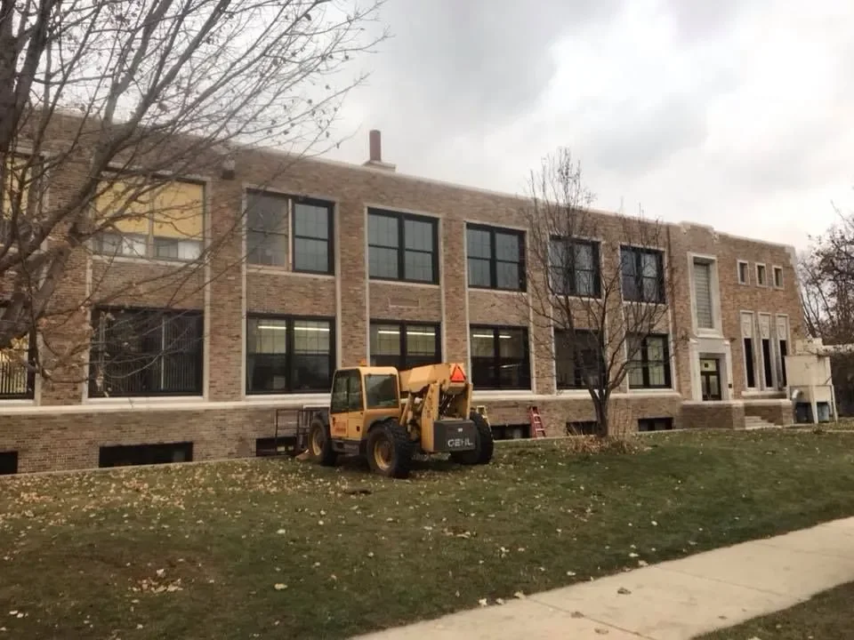 A yellow construction vehicle parked on a grassy area in front of a multi-story brick building under possibly overcast skies.