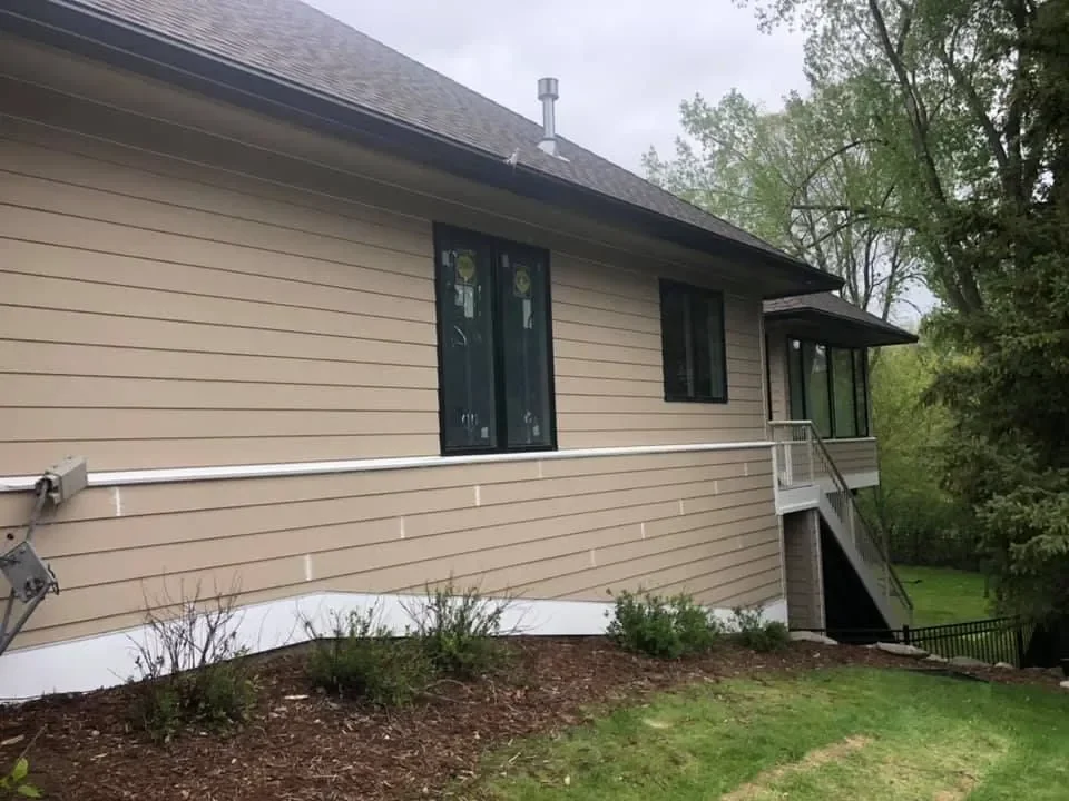 The back of a beige house with vinyl siding, a small deck with stairs, and a well-maintained lawn with bushes.