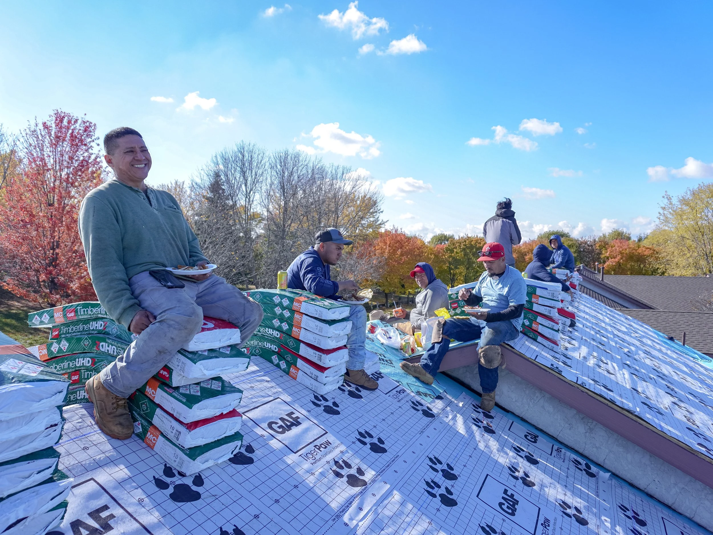 Group of people sitting and standing on a roof during fall, surrounded by colorful trees, with blue sky and clouds overhead