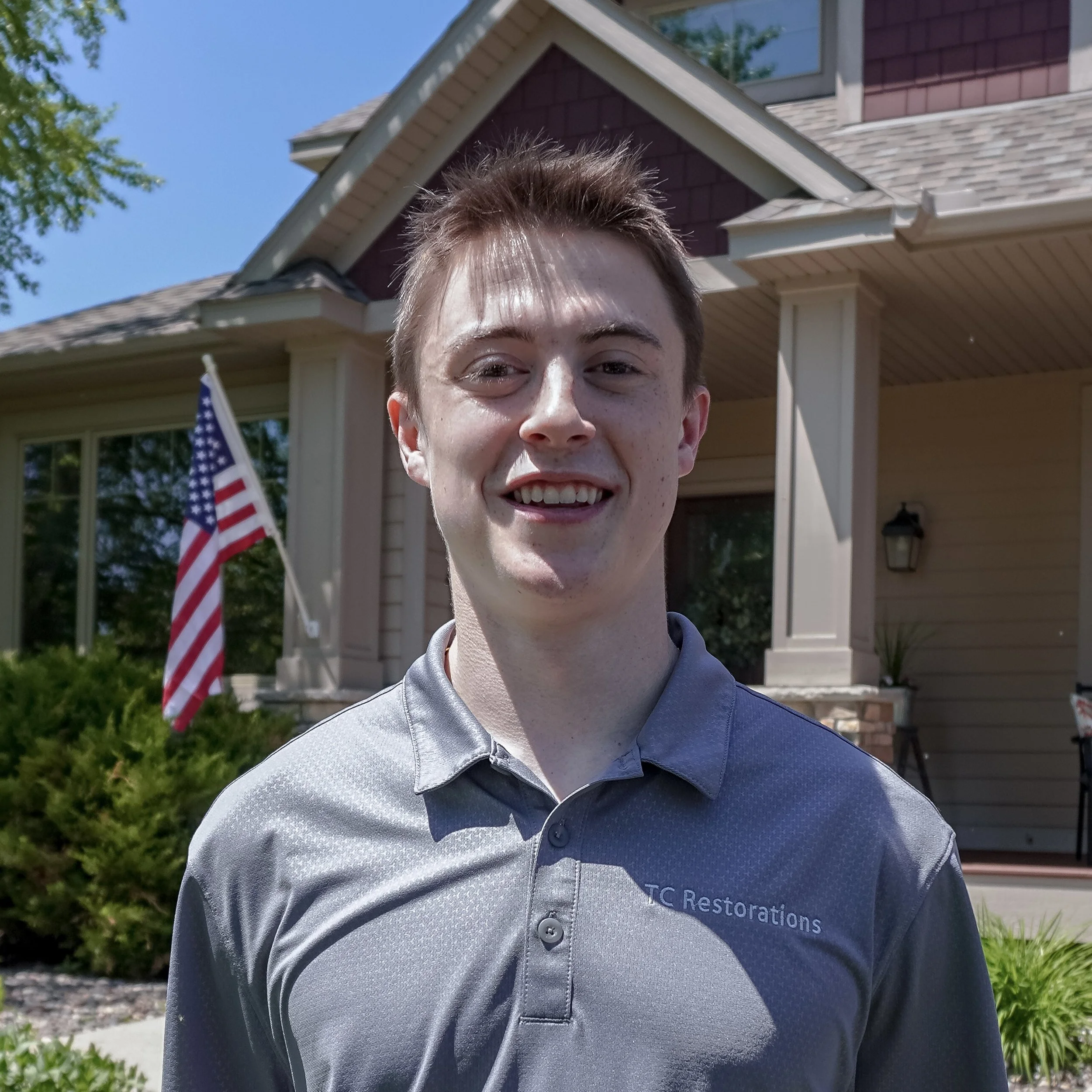 A young man smiling outdoors in front of a house with a porch, American flags, and greenery.