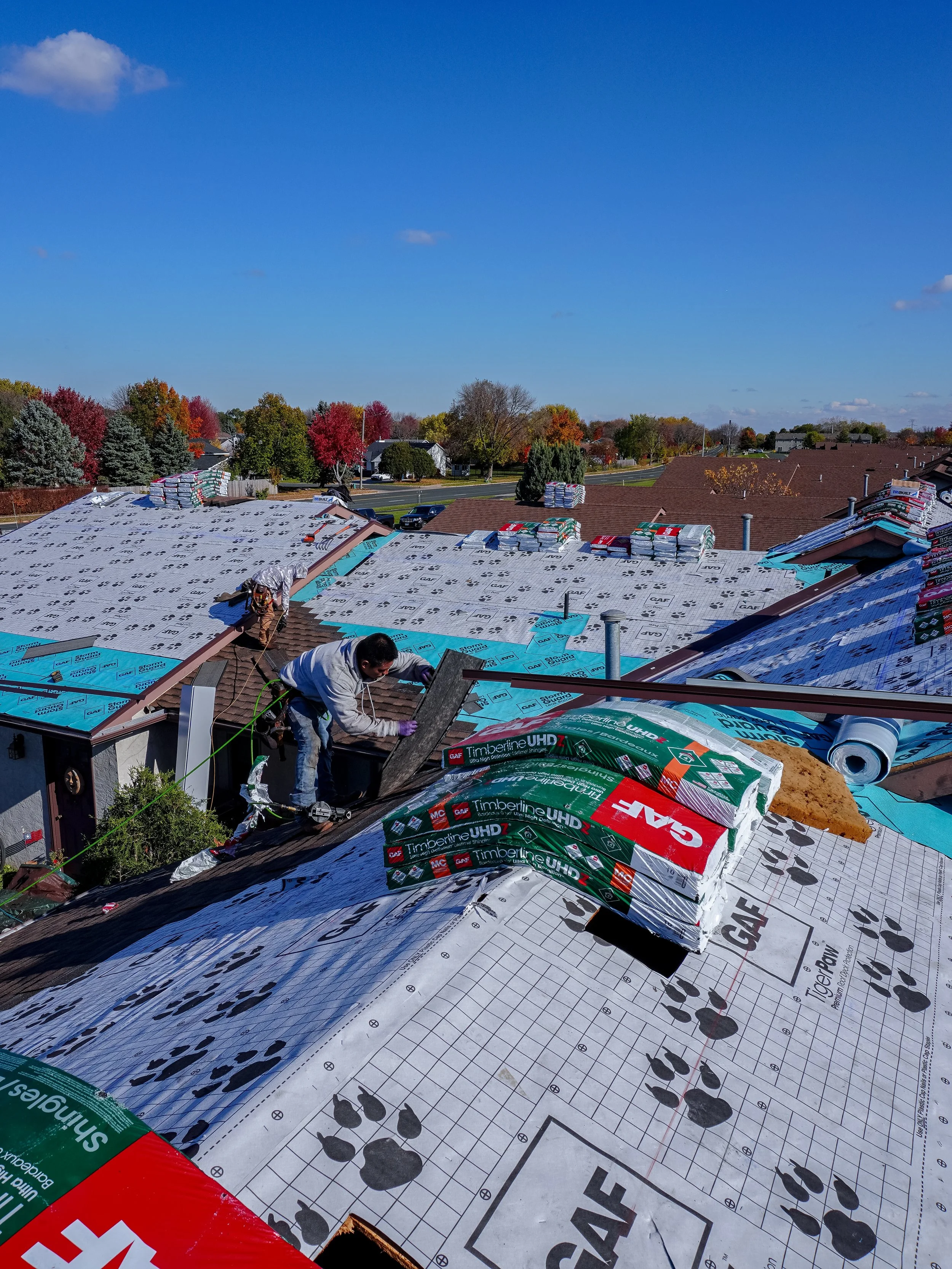 Roof under construction with workers installing shingles, stacks of roofing materials, and a clear blue sky in the background.