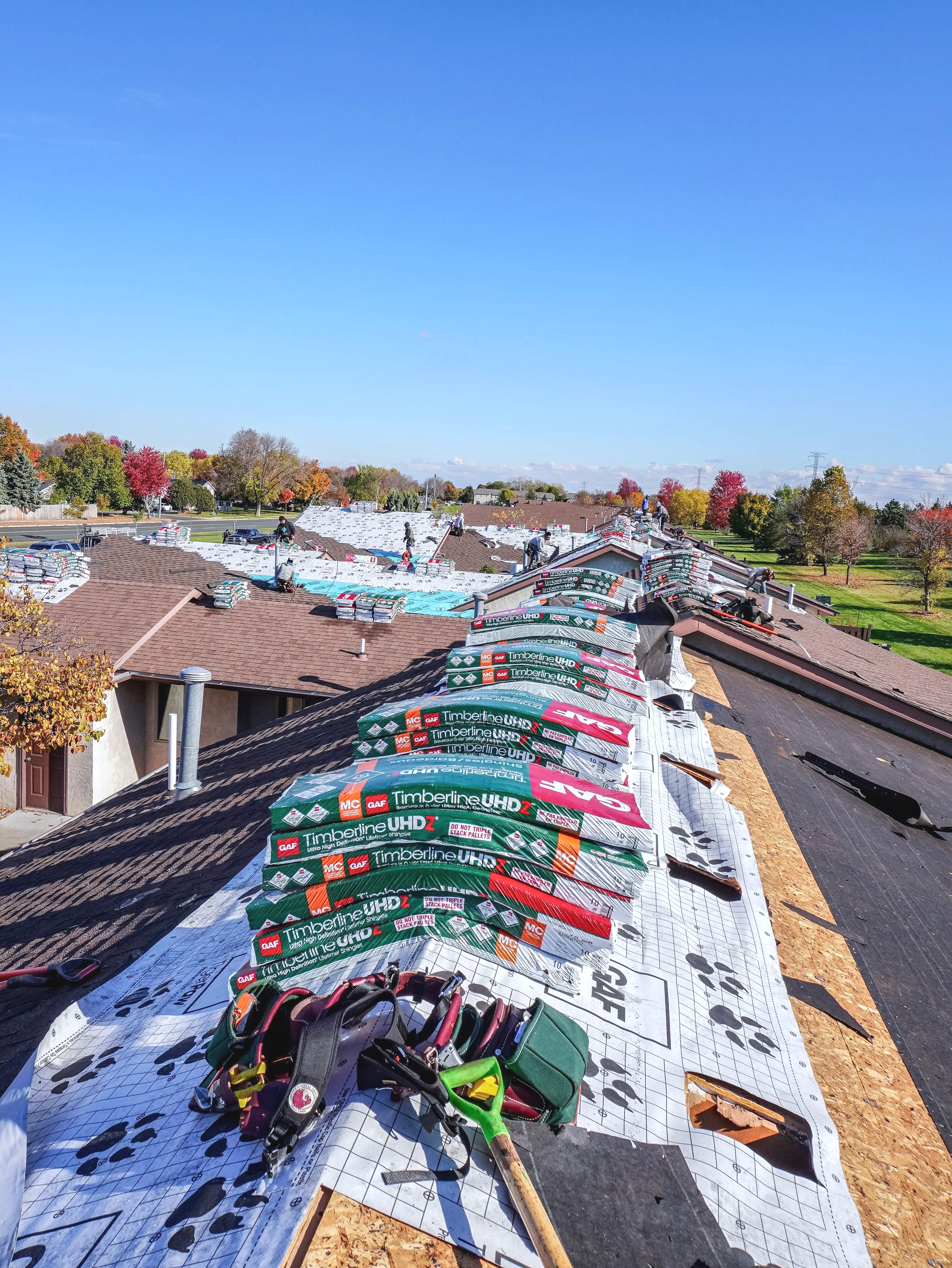 Roofing installation under construction, workers installing new roof shingles, tools and building materials on roof, trees with fall foliage in background, clear blue sky.