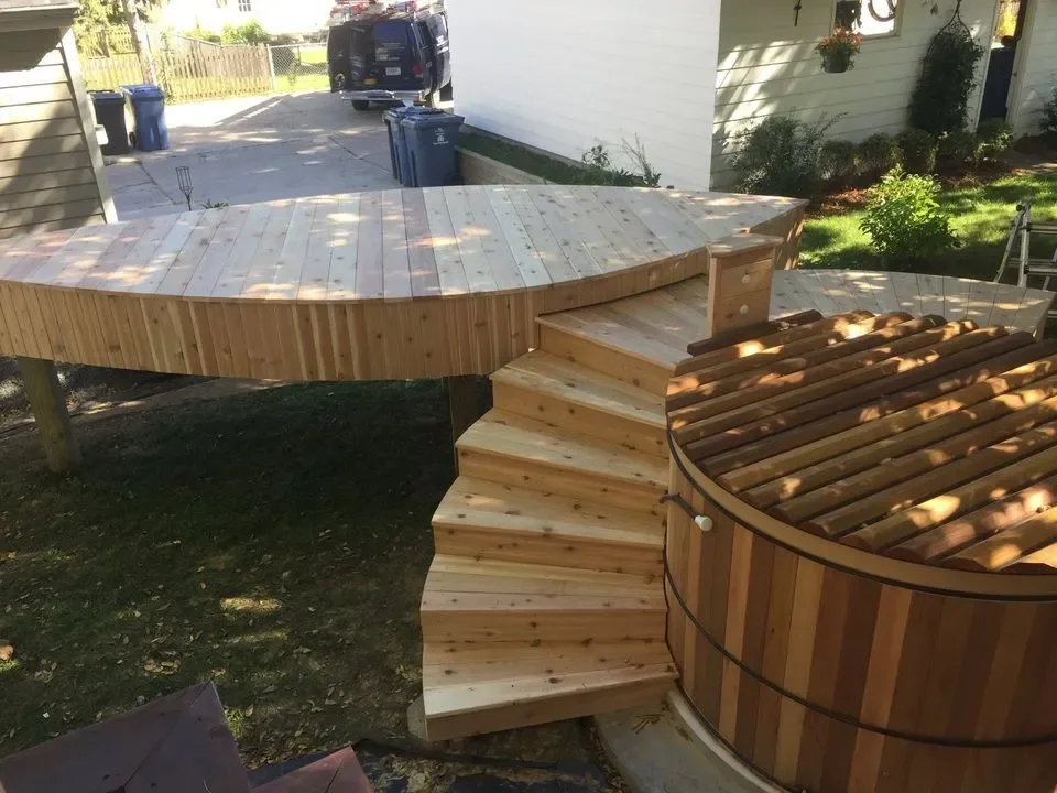 A wooden deck with curved stairs leading up to a house porch, surrounded by a backyard with grass and plants.