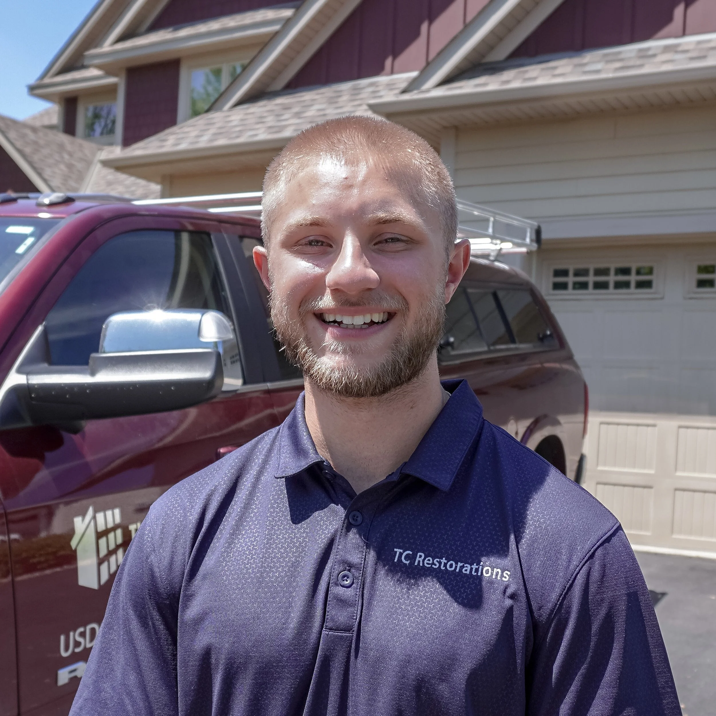 Close-up of a smiling young man with short blond hair and a beard, standing outdoors with a house and a red pickup truck in the background.