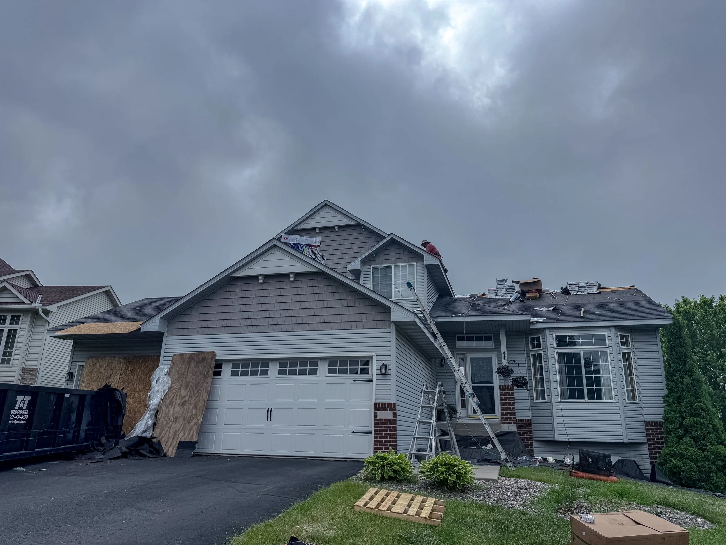 The image shows a two-story house with a gray exterior and white trim. A worker is on the roof, possibly performing repairs or inspection, with roofing materials around. There is a ladder leaning against the house, a trash bin nearby, and some plywood and cardboard on the ground. The sky is overcast with dark clouds, suggesting stormy weather.