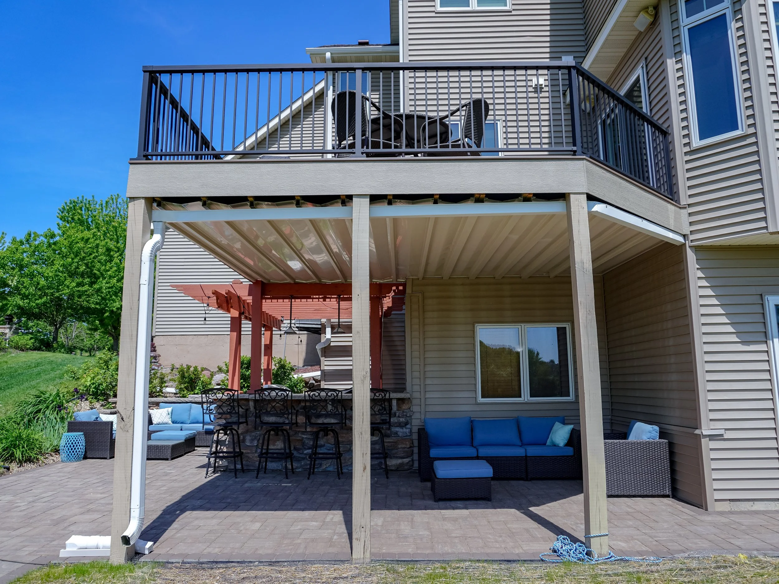 A backyard patio with furniture underneath a deck, with a house in the background and a tree to the left.