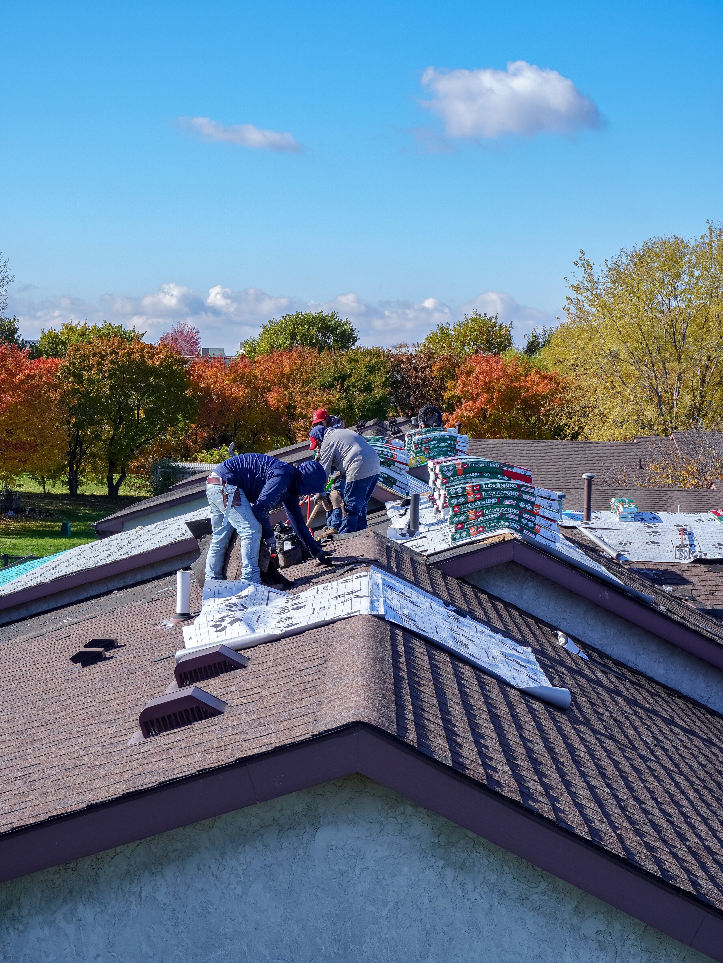 Workers repair a roof with shingles and roofing materials in a suburban neighborhood with trees and a blue sky.