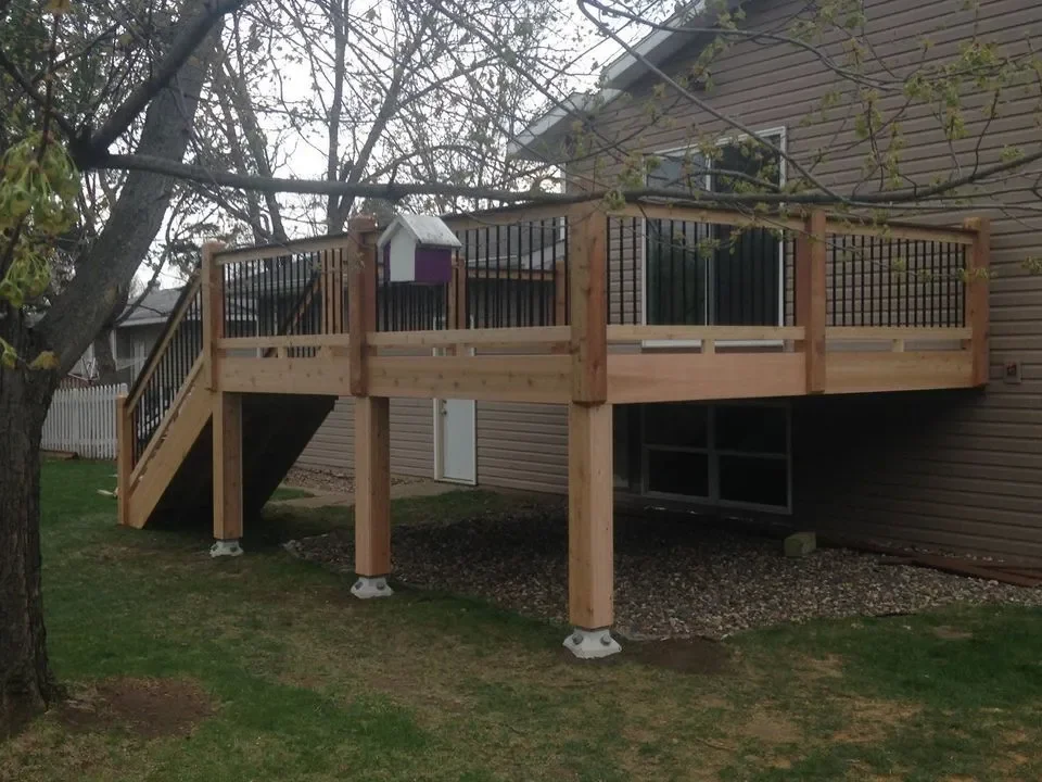 Wooden deck with black railings attached to a house in a backyard. The deck has stairs leading down to the grass, and a tree with branches is in the foreground.