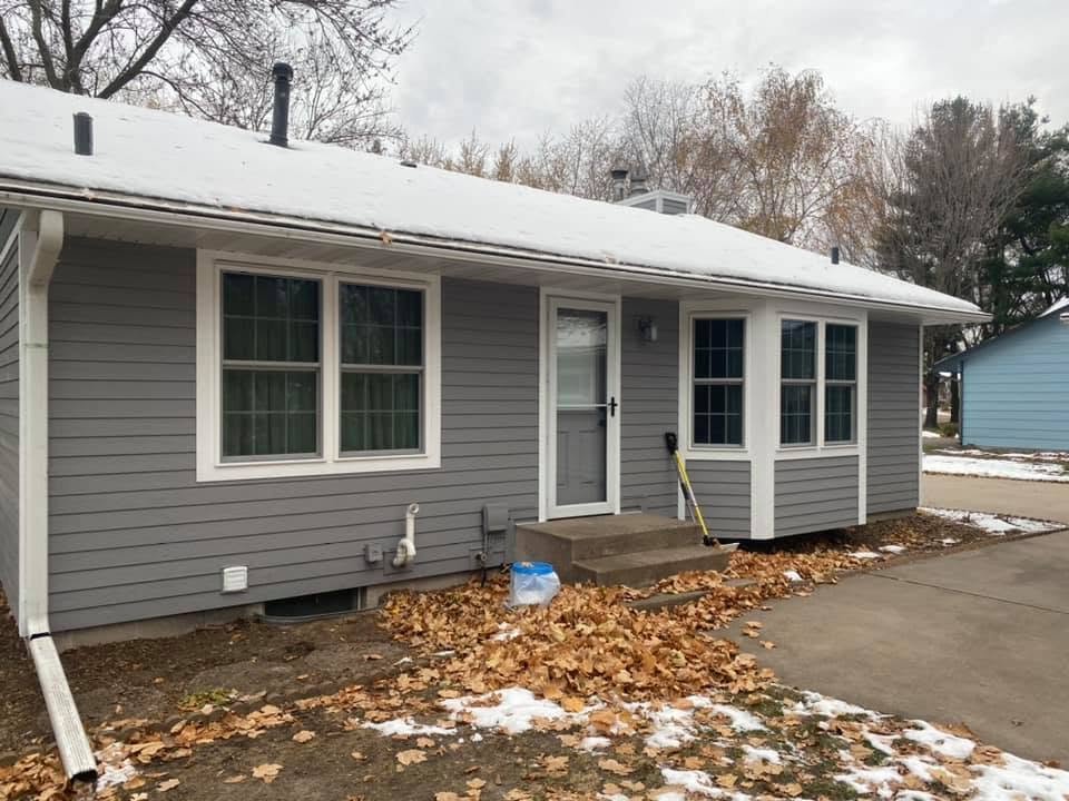 A gray house with a snow-covered roof and two large windows. There is a small set of stairs leading to the back door, which has a porch light above. Fallen leaves are on the ground, and a yellow-handled shovel is leaning against the house near the do