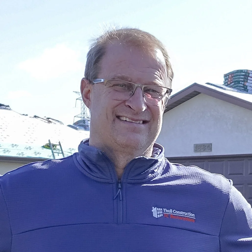 A man with glasses and short hair smiling outdoors in front of a house with a garage, wearing a blue jacket with 'Thull Construction' logo.