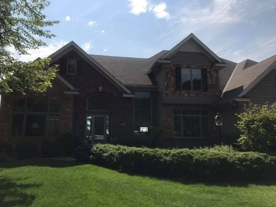 A two-story brick house with a gabled roof, front yard with green grass and shrubs, and partially cloudy sky.