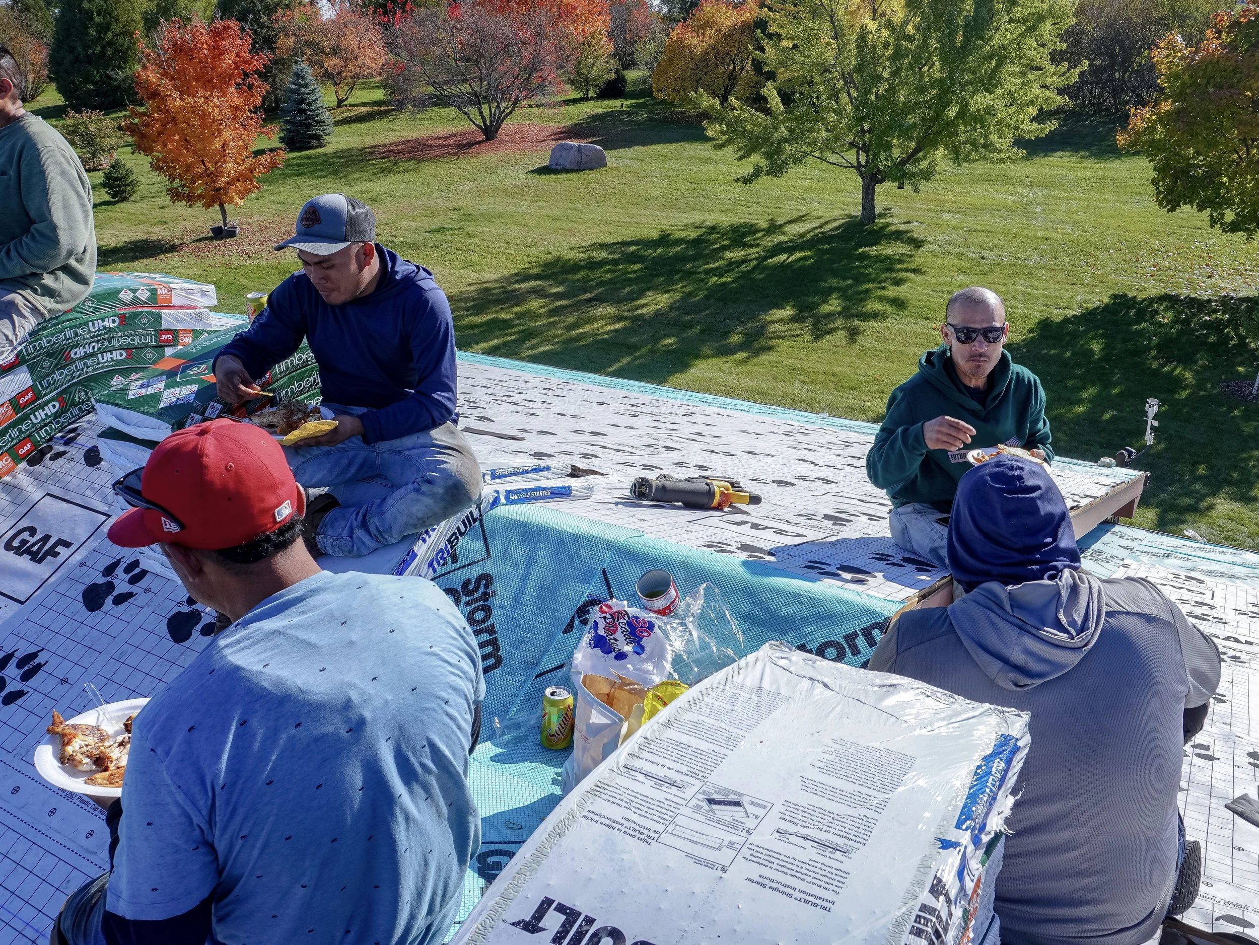 People sitting on a roof-top with food, drinks, and supplies, having a picnic or outdoor gathering in a park during fall, with trees with colorful autumn leaves in the background.