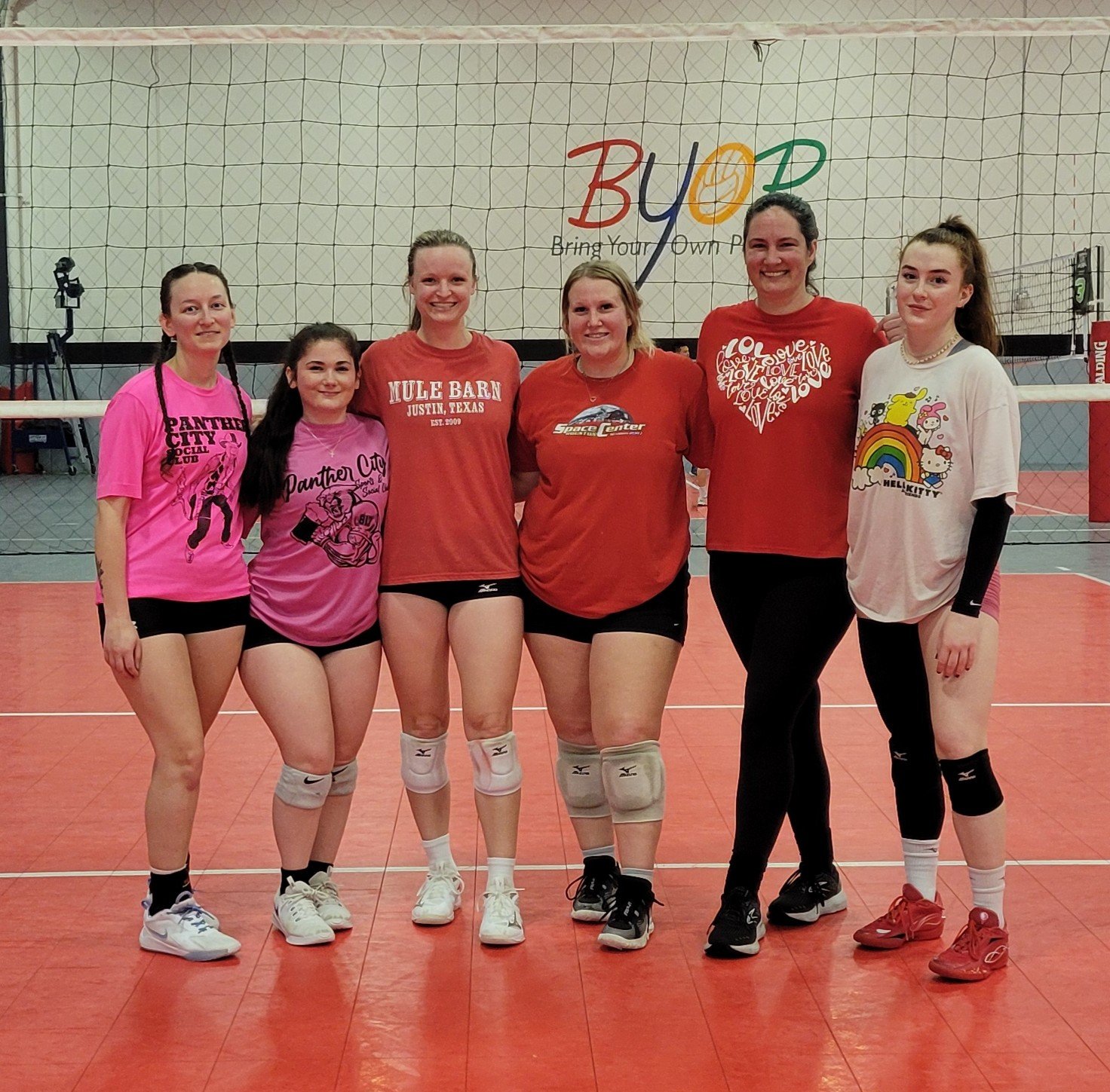 Six women stand together on an indoor volleyball court, smiling at the camera. They wear sports attire, including knee pads, with a volleyball net and a colorful gym wall in the background.