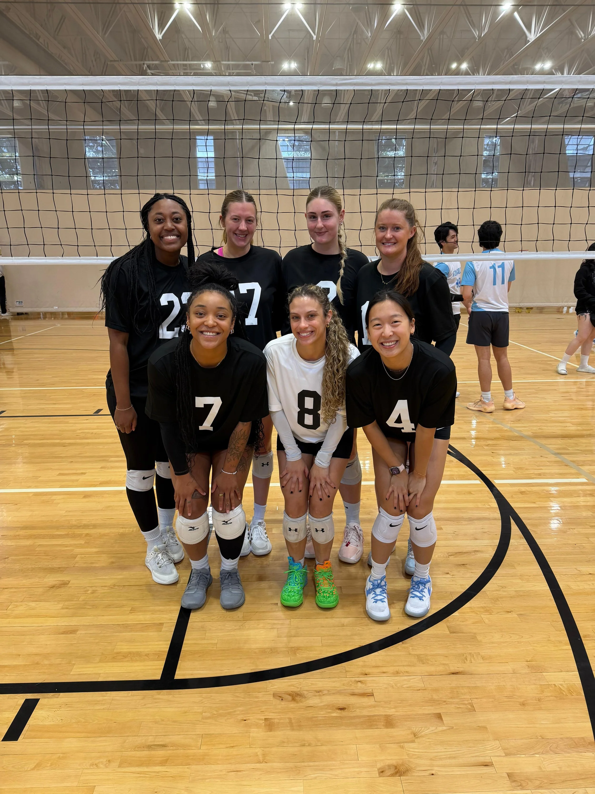 Seven young women volleyball players in black jerseys with white numbers, smiling and posing for a photo on an indoor volleyball court. Two women in the background are wearing light blue and white volleyball uniforms.