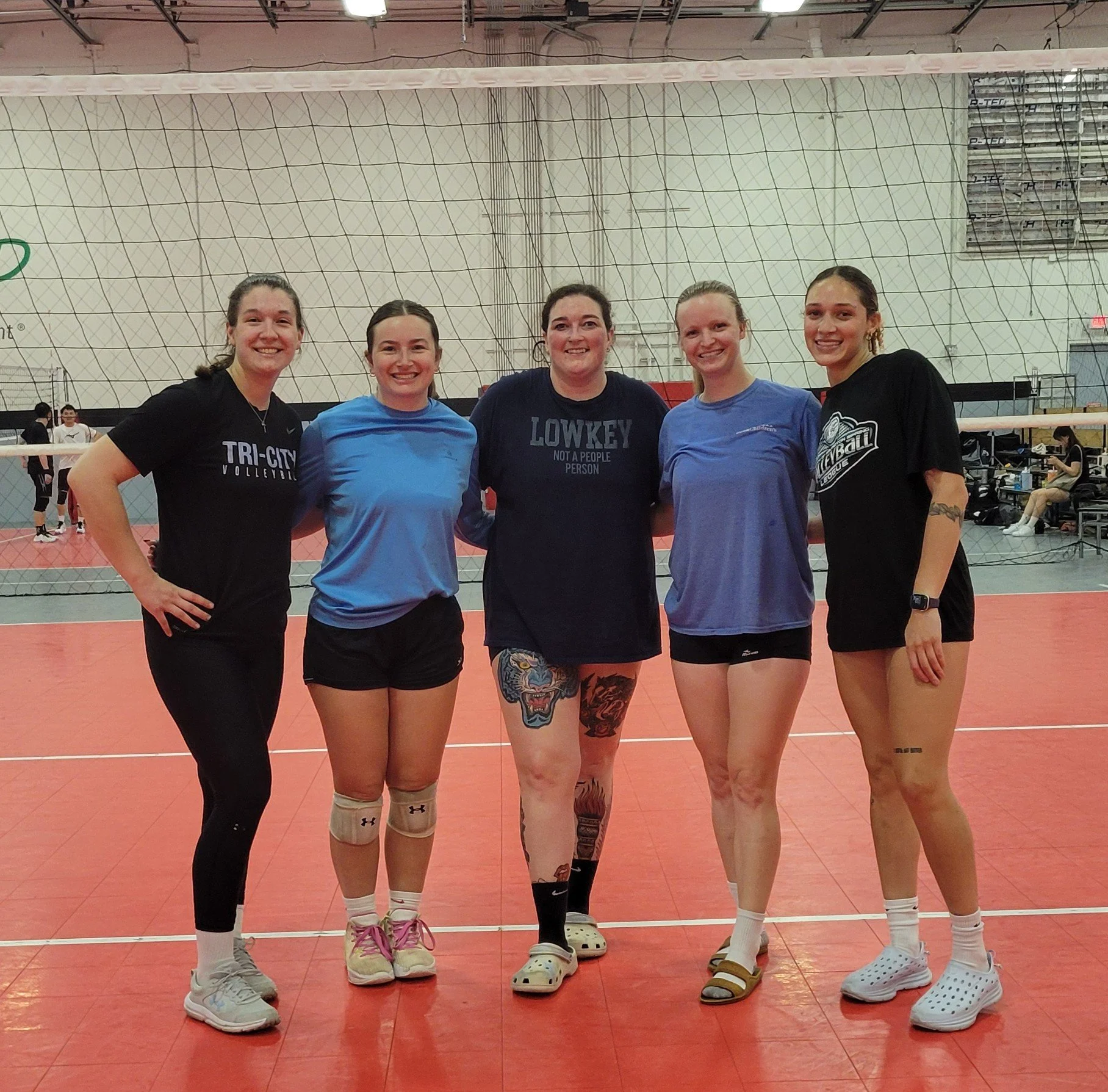 Group of five young women standing together on an indoor volleyball court. They are dressed in athletic clothing, with some wearing volleyball shirts, shorts, and socks, and others in casual sportswear. They are smiling and standing in front of a volleyball net, with a gymnasium, other players, and spectators visible in the background.