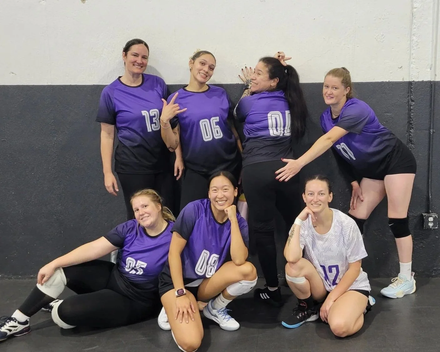 A group of seven women, dressed in purple volleyball jerseys with various numbers, posing together against a two-tone wall in an indoor gym. They are smiling and making playful gestures.