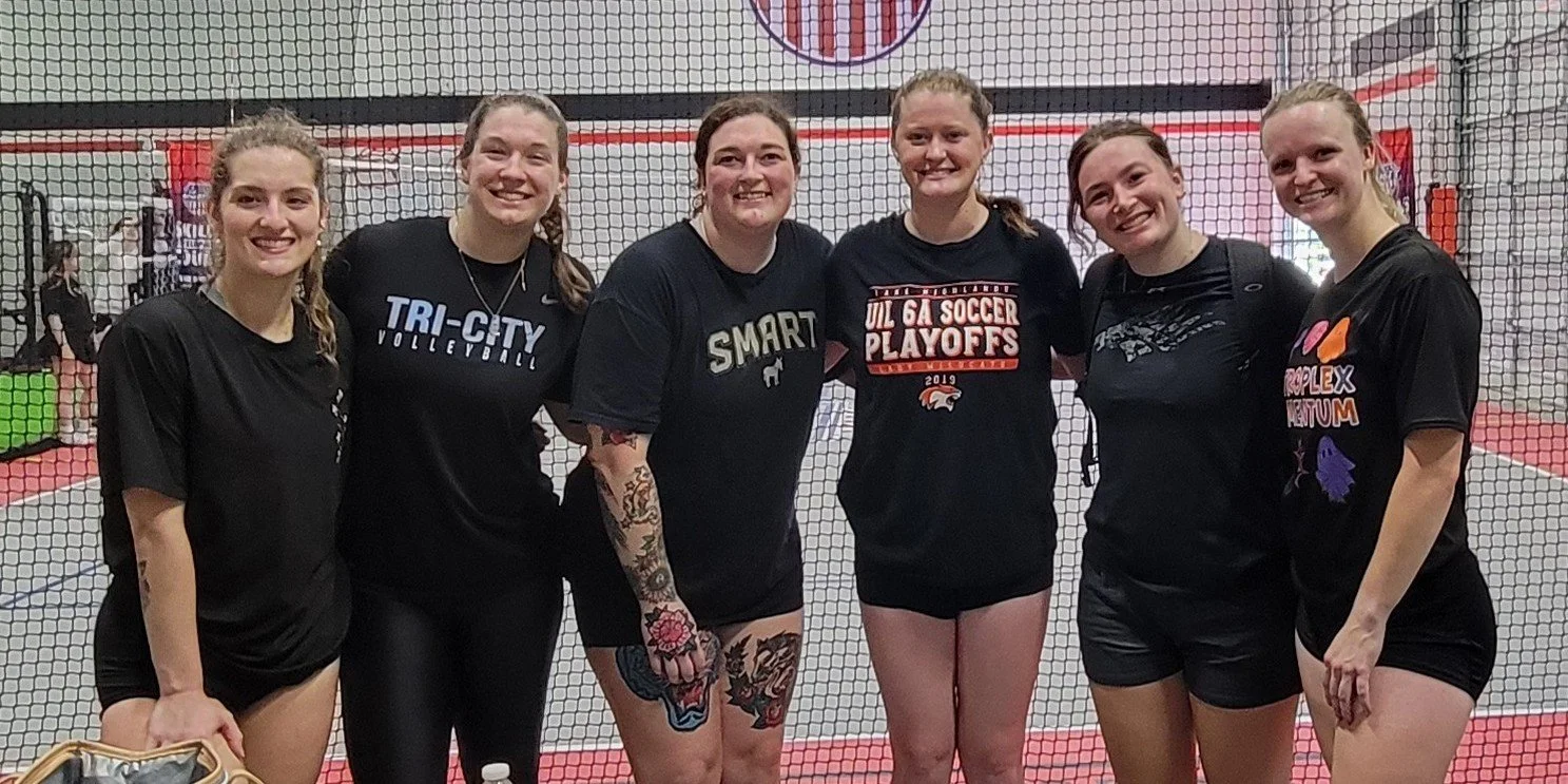 Six women in black athletic shirts standing together inside a volleyball gym.