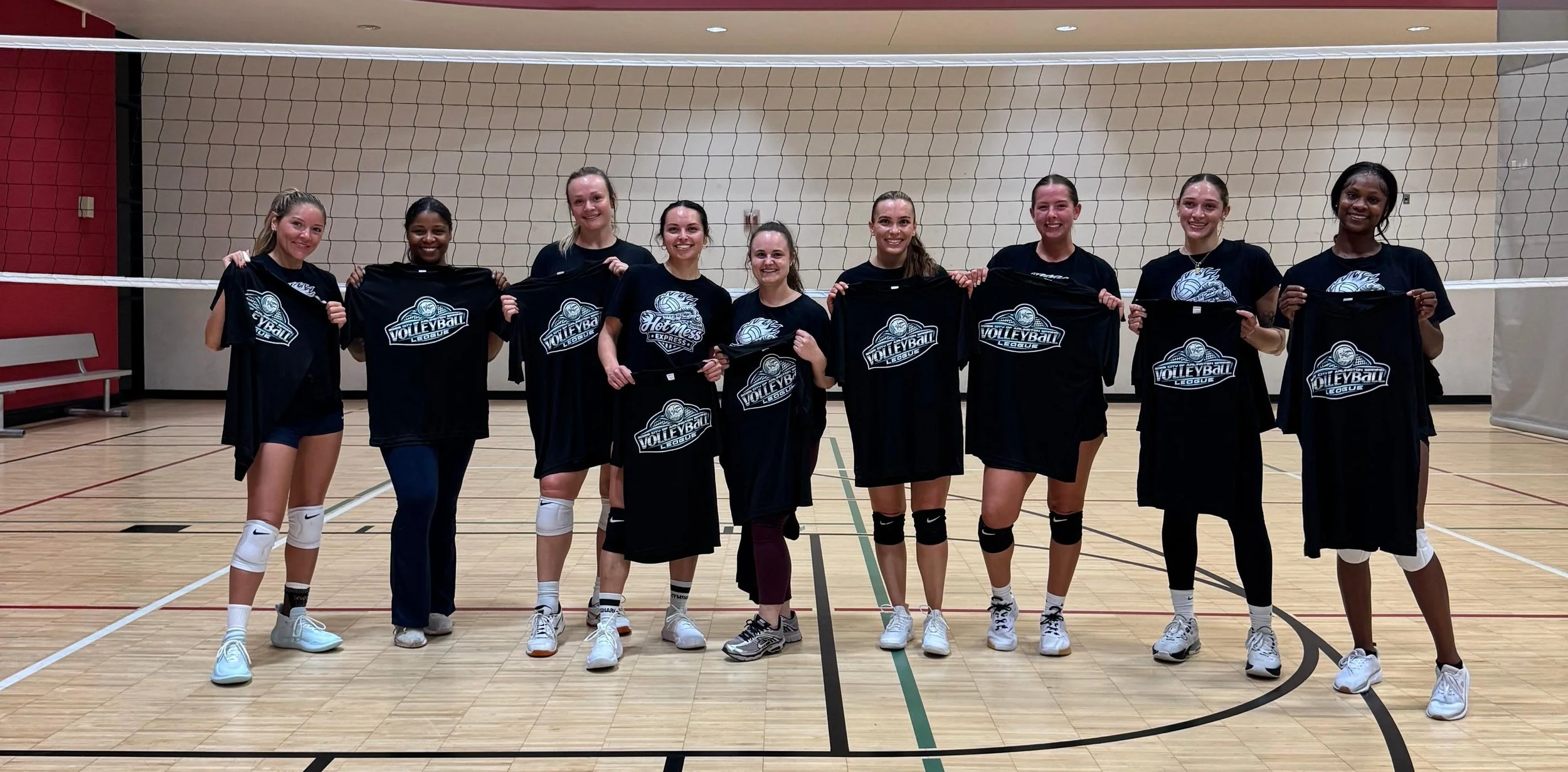 A group of nine female volleyball players standing in a gymnasium, holding black T-shirts with a volleyball logo and the words 'Volleyball League' and 'Hot Mess Express.' They are smiling and wearing athletic gear including knee pads and sneakers.