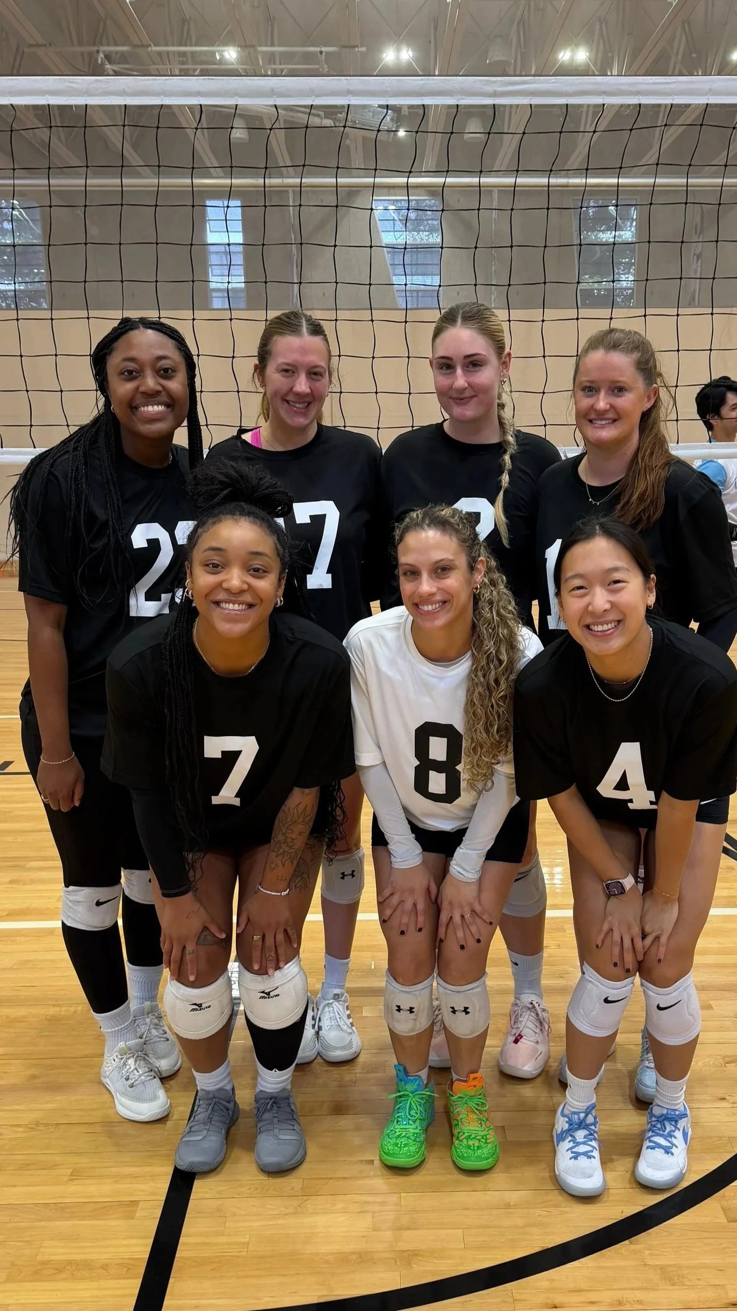 Group of nine women volleyball players in uniform posing behind a net in a gymnasium.