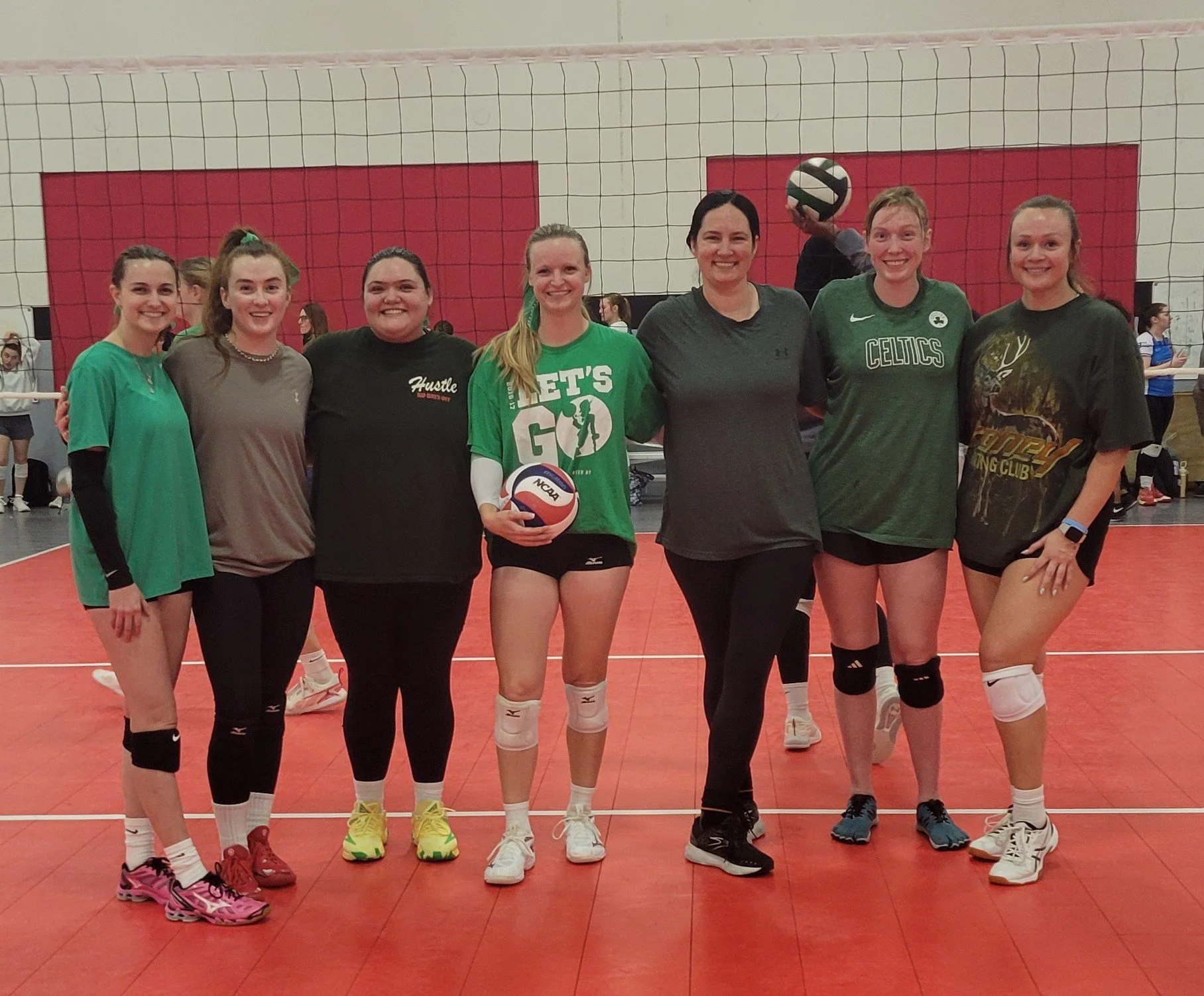 A group of seven women standing together on an indoor volleyball court posing for a photo. They are smiling and dressed in athletic clothing, with a volleyball net in the background.